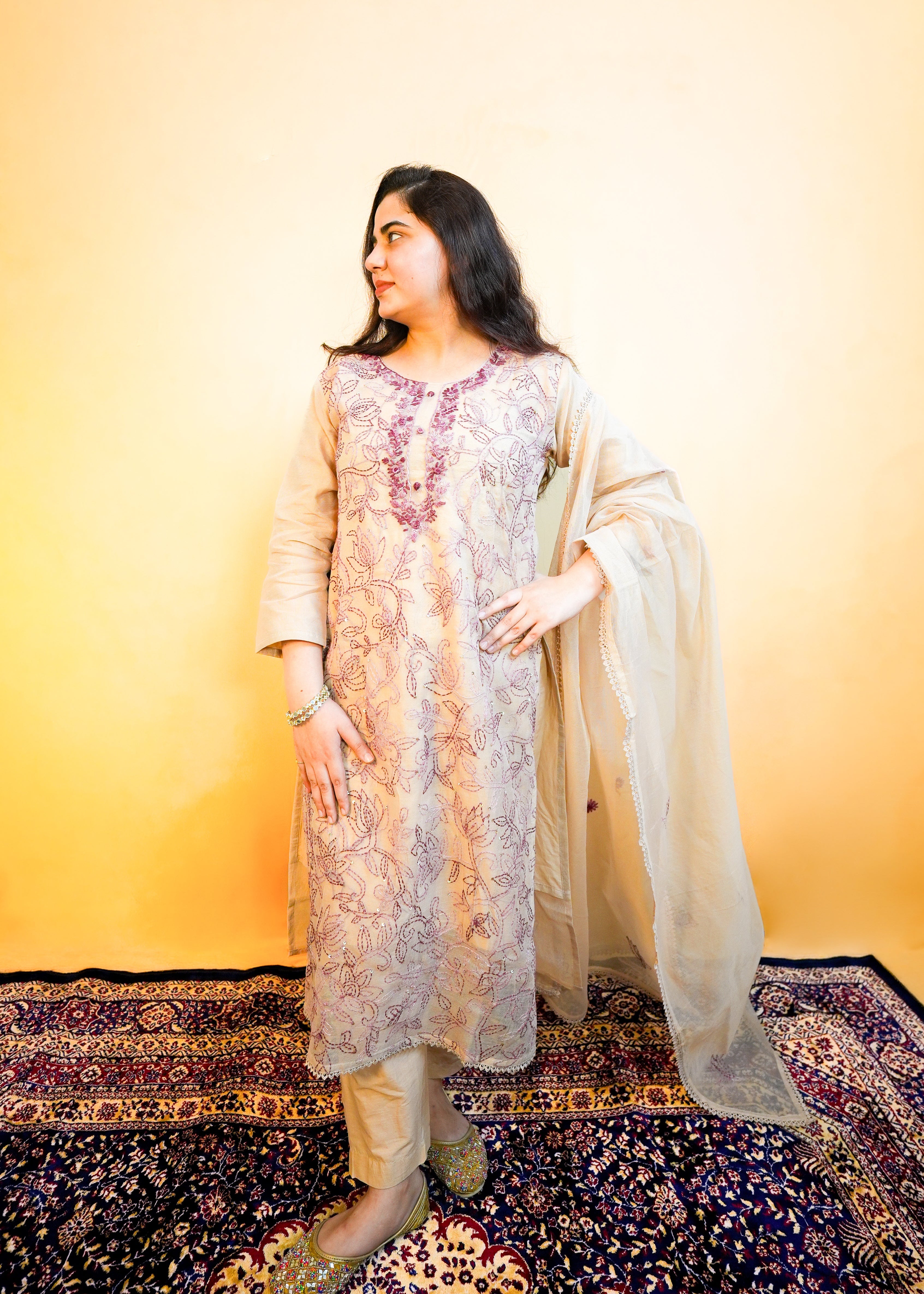 Woman in traditional outfit standing on a patterned rug with a yellow wall background