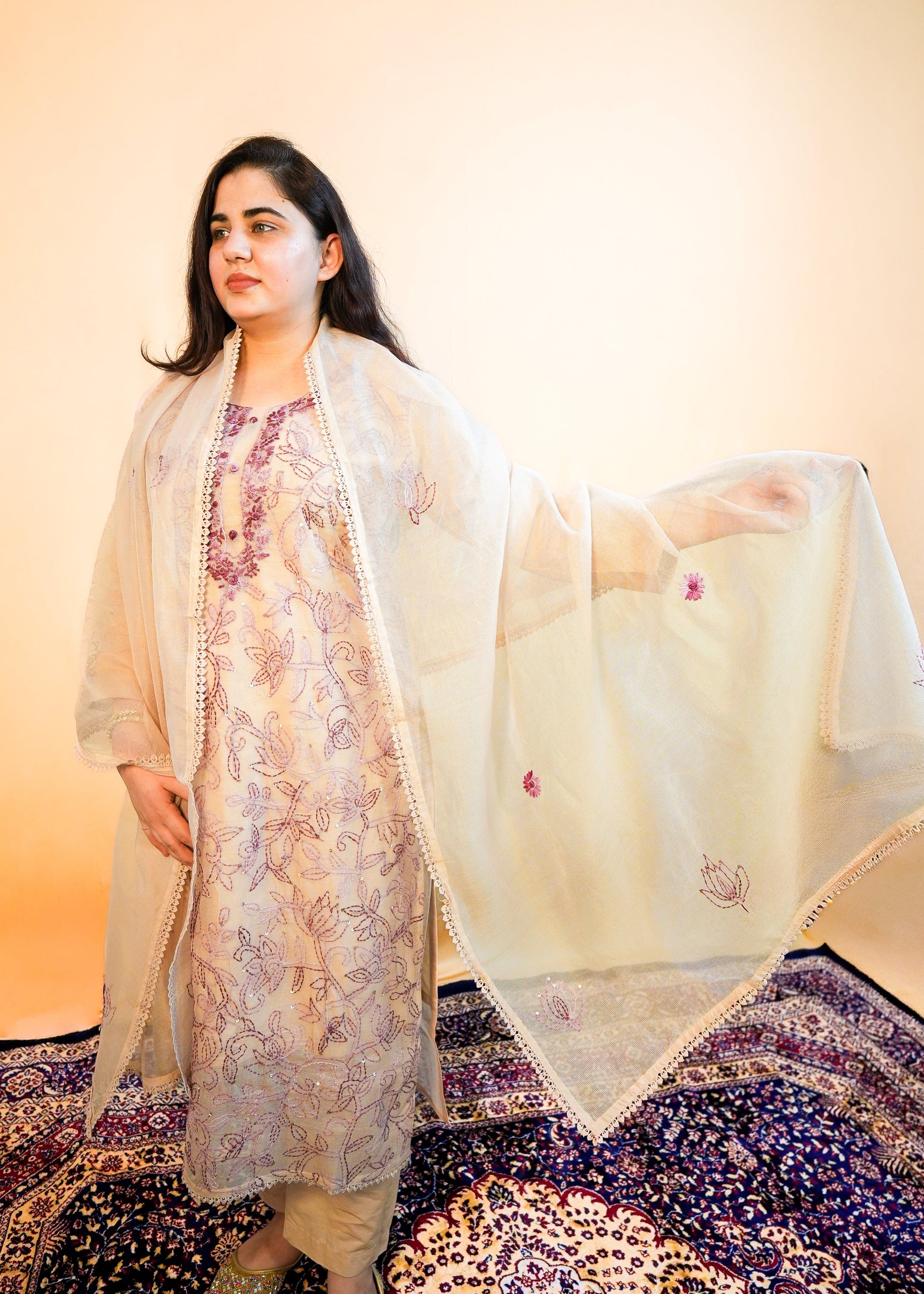Woman in traditional attire standing on a patterned rug with a plain background