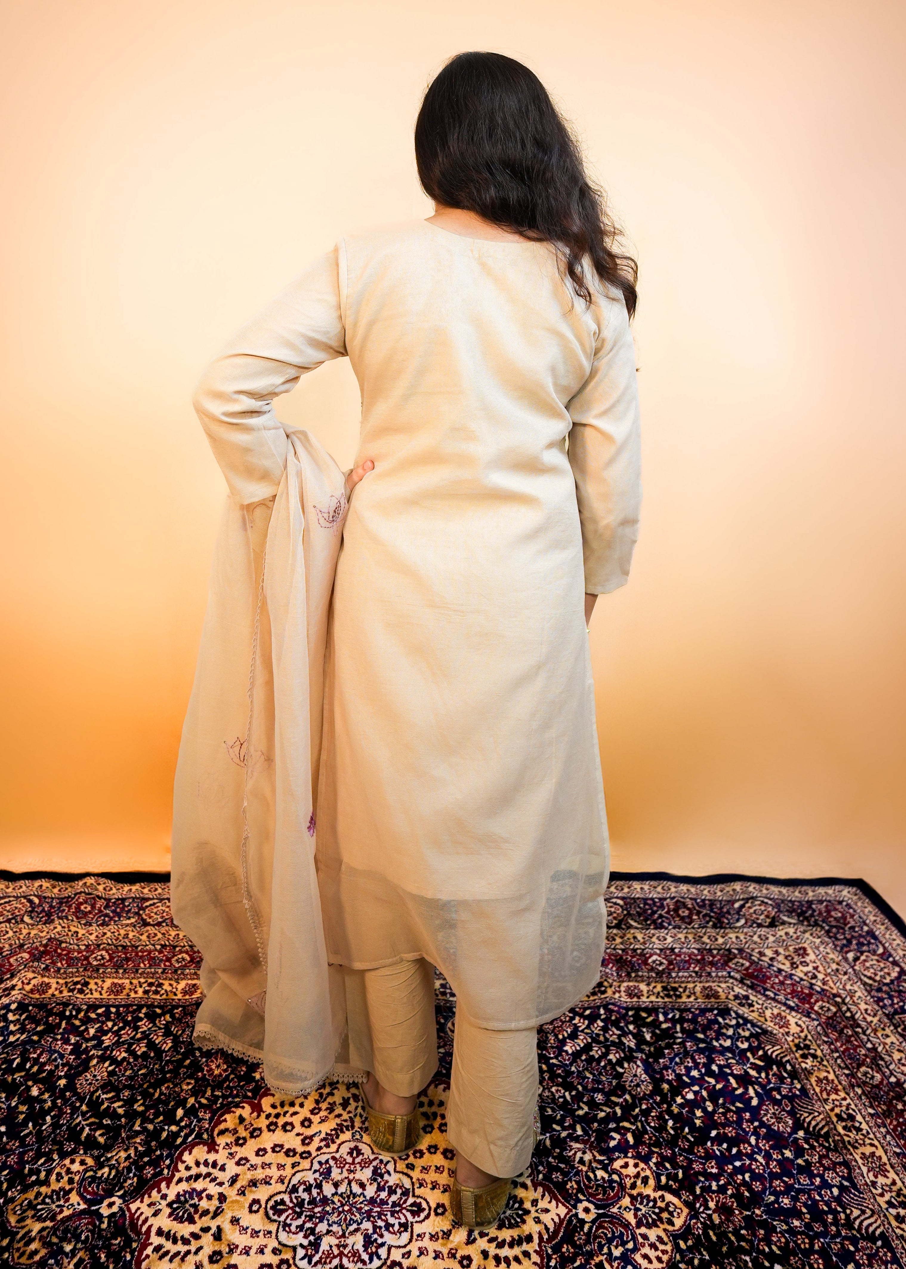 Person wearing a long cream-colored dress standing on a patterned rug with a warm-toned wall in the background.