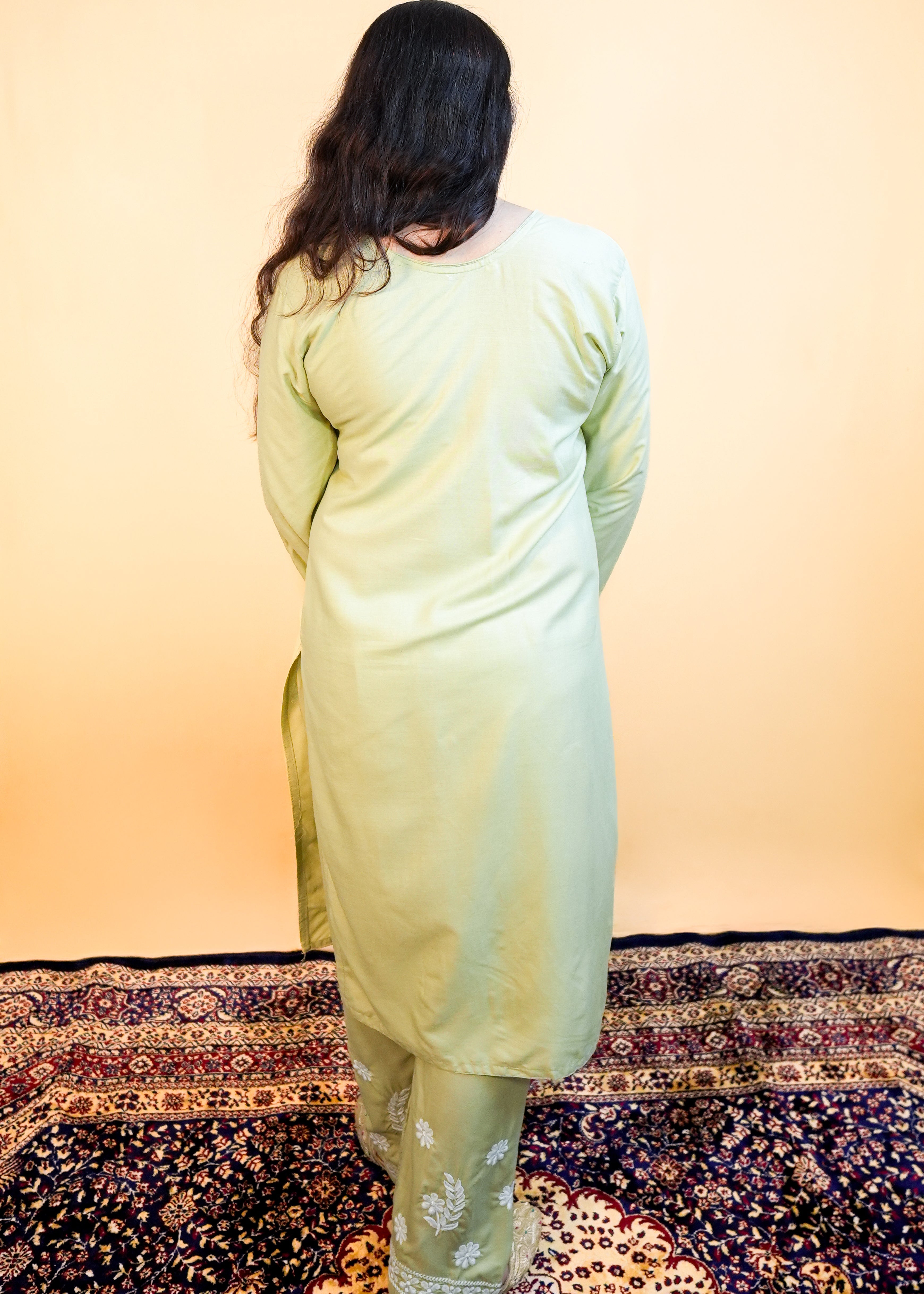 Person wearing a light green traditional outfit standing on a patterned rug with a gradient wall.