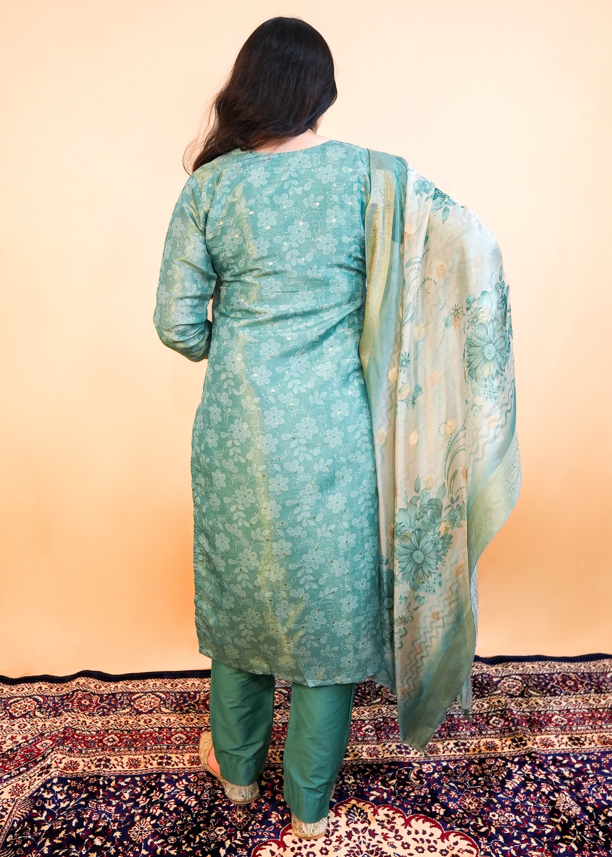 Person wearing a teal traditional outfit with a patterned shawl on a decorative rug.