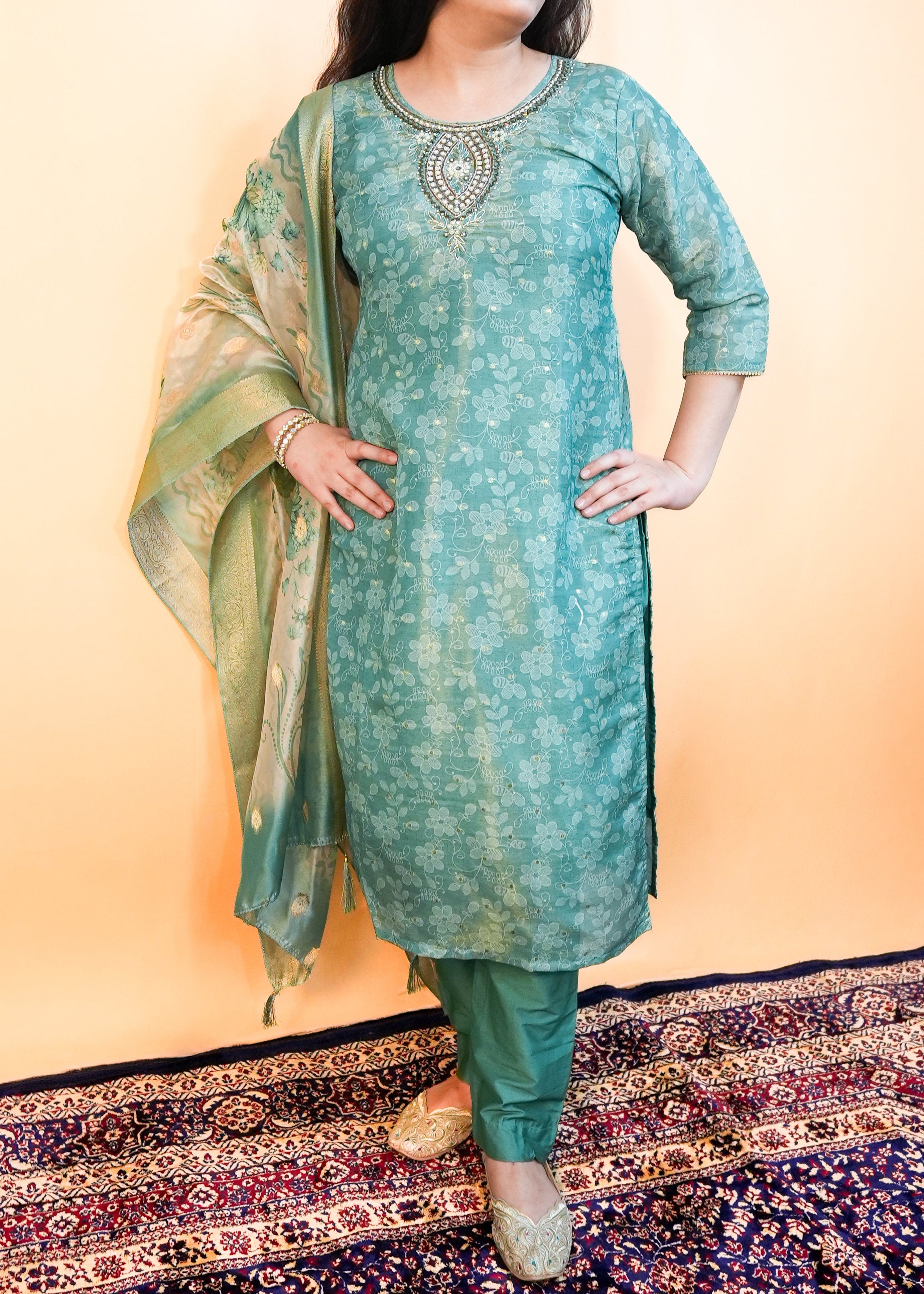 Woman wearing a teal traditional outfit with a patterned rug below