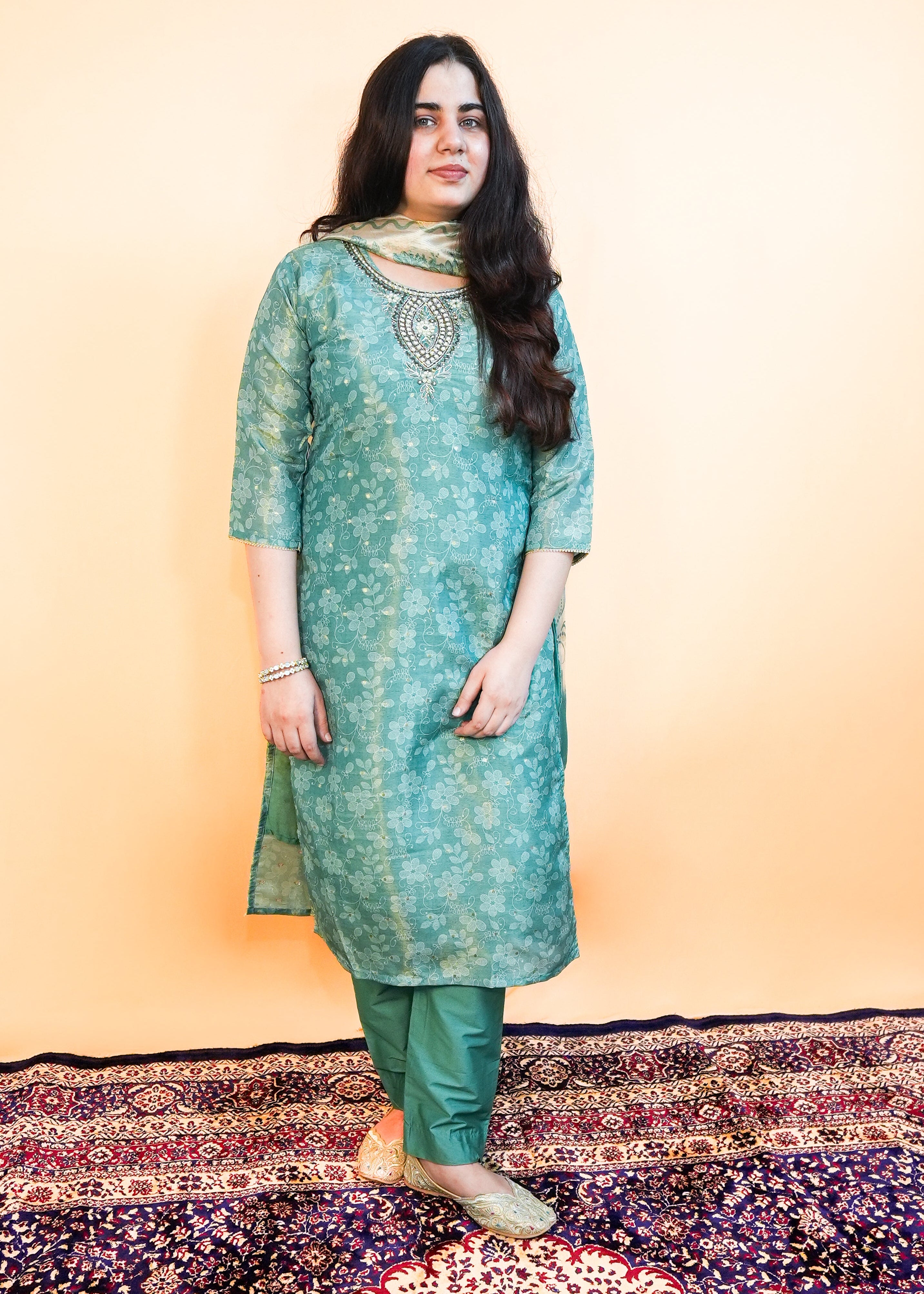 Woman wearing a green patterned kurta standing on a decorative rug with a plain background
