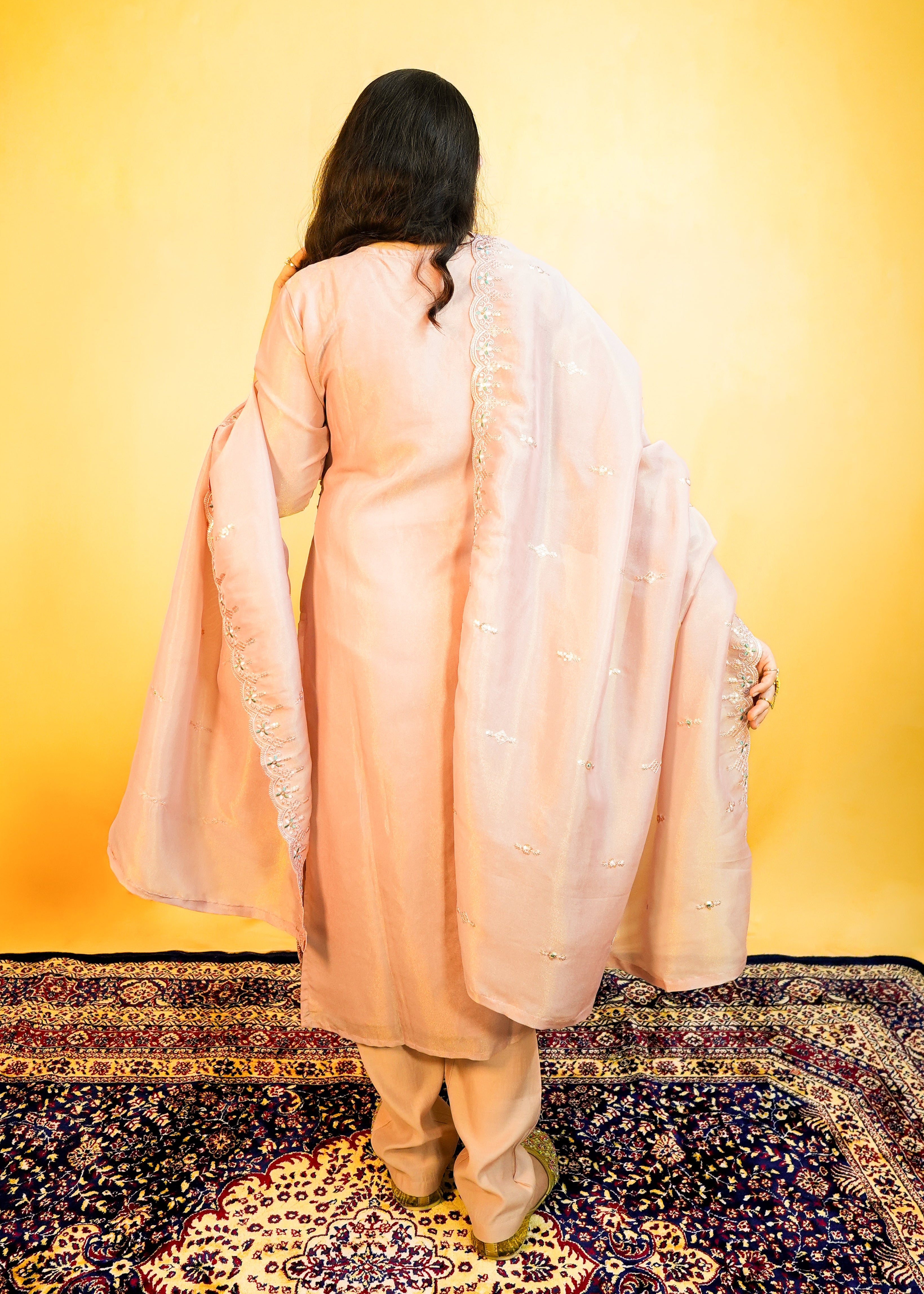 Person wearing a light pink traditional outfit standing on a patterned rug with a yellow background