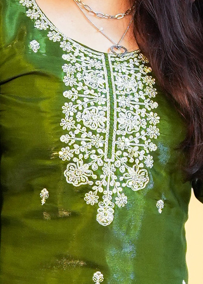 Woman in a green traditional outfit with white patterns standing on a patterned rug against a yellow wall.