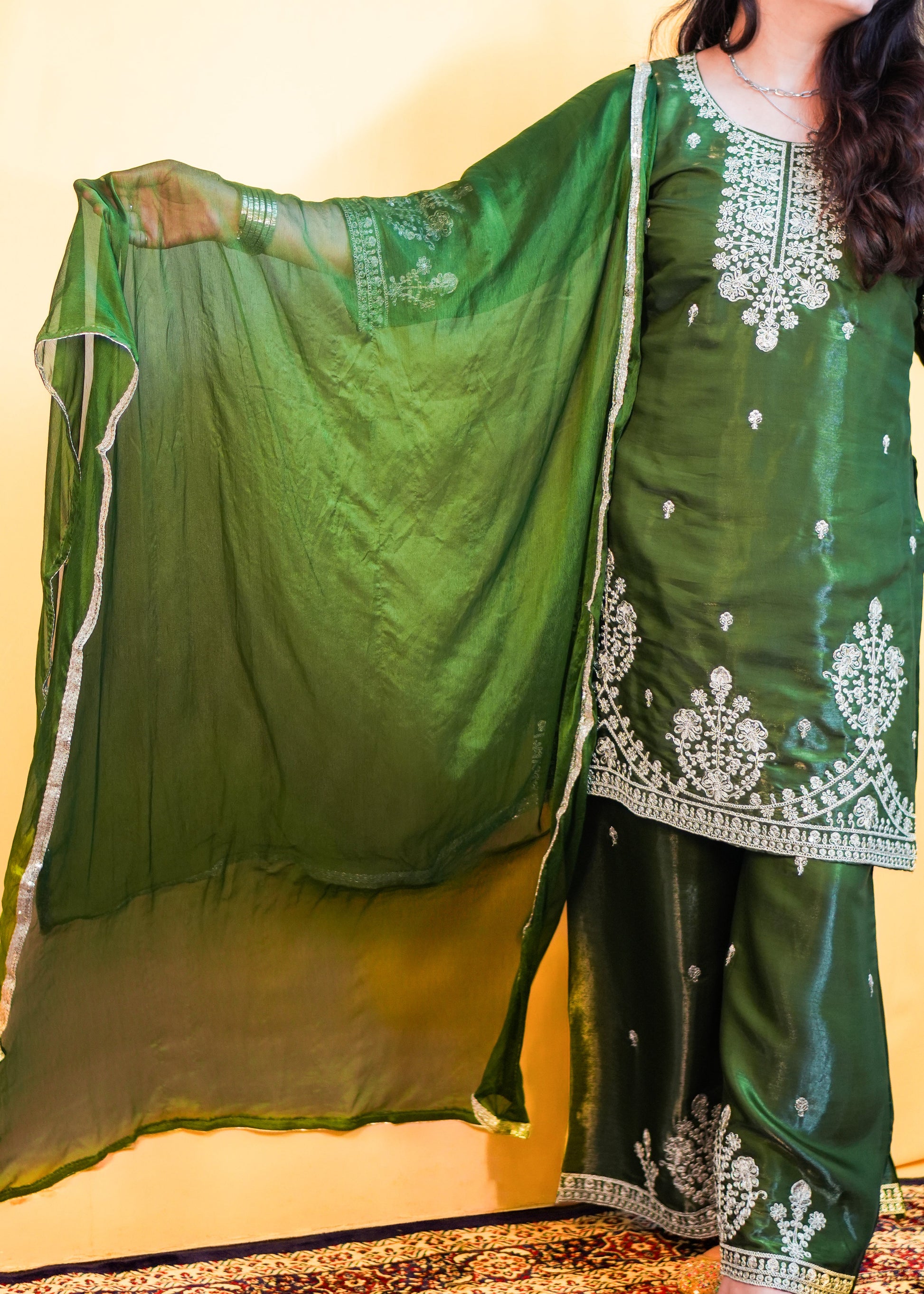 Woman in a green traditional outfit with white embroidery standing on a patterned rug.
