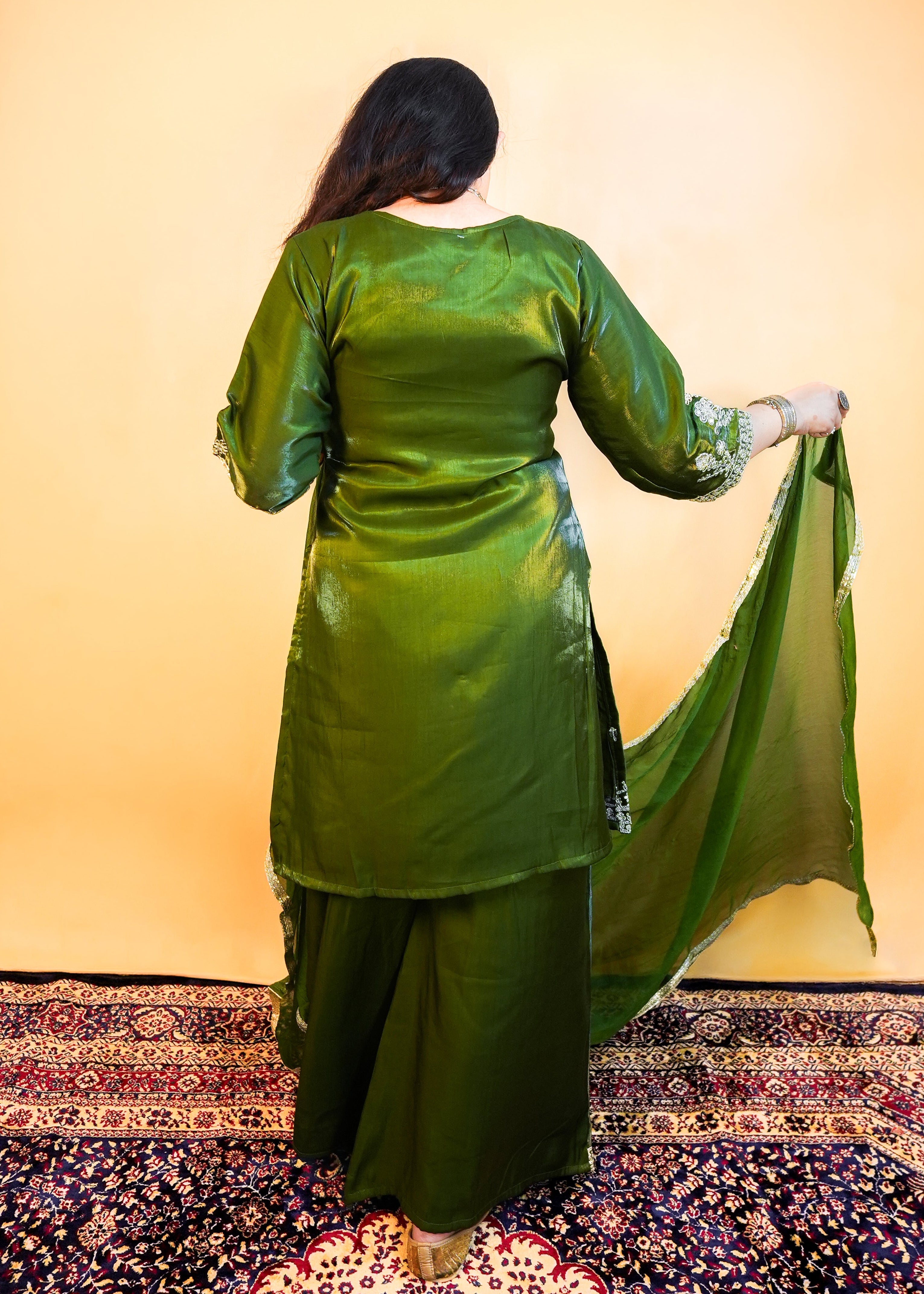 Woman in a green traditional outfit standing on a patterned rug with a warm-toned background