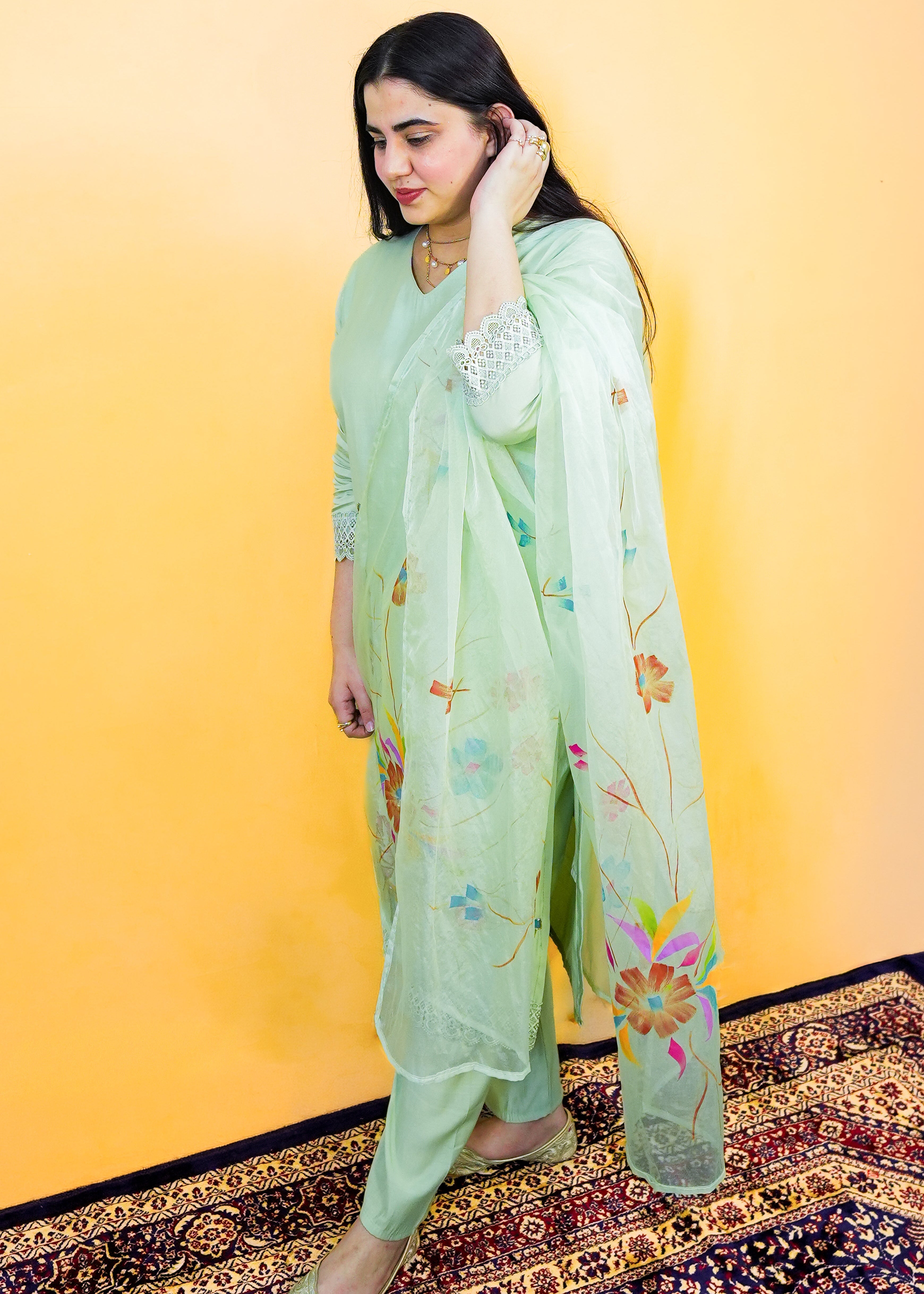 Woman in a light green traditional outfit with floral patterns against a yellow wall.
