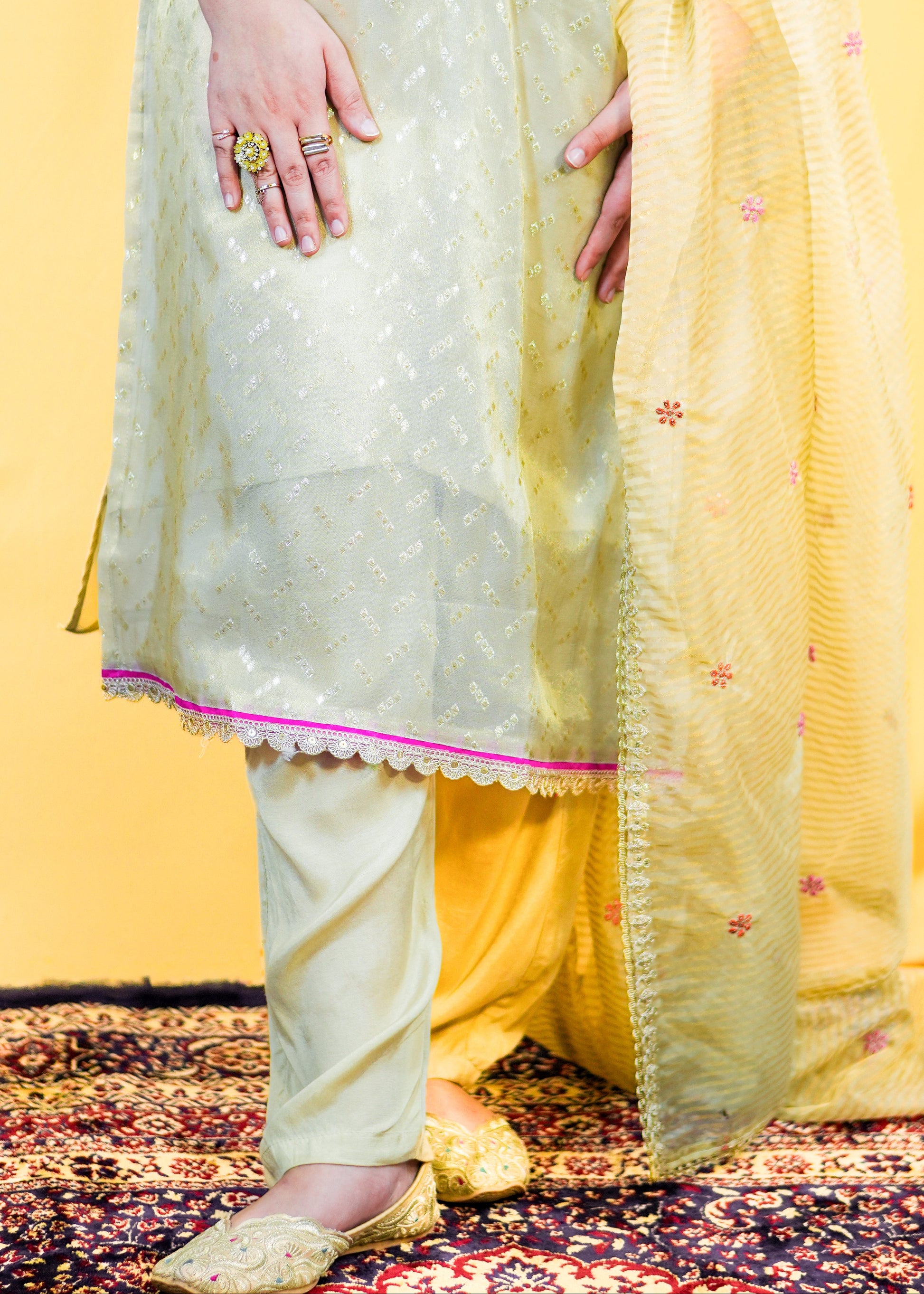 Person wearing a light green traditional outfit with a yellow dupatta on a patterned rug.