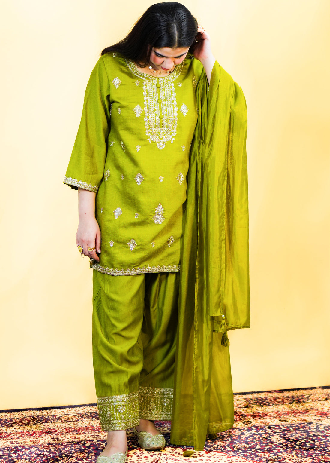 Woman wearing a green traditional outfit with embroidery on a patterned rug.