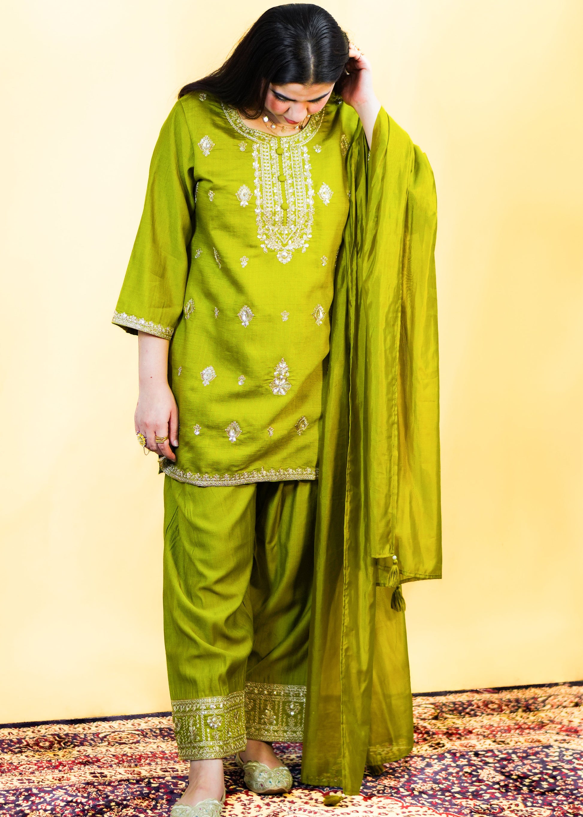 Woman wearing a green traditional outfit with embroidery on a patterned rug.