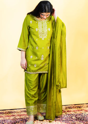 Woman wearing a green traditional outfit with embroidery on a patterned rug.