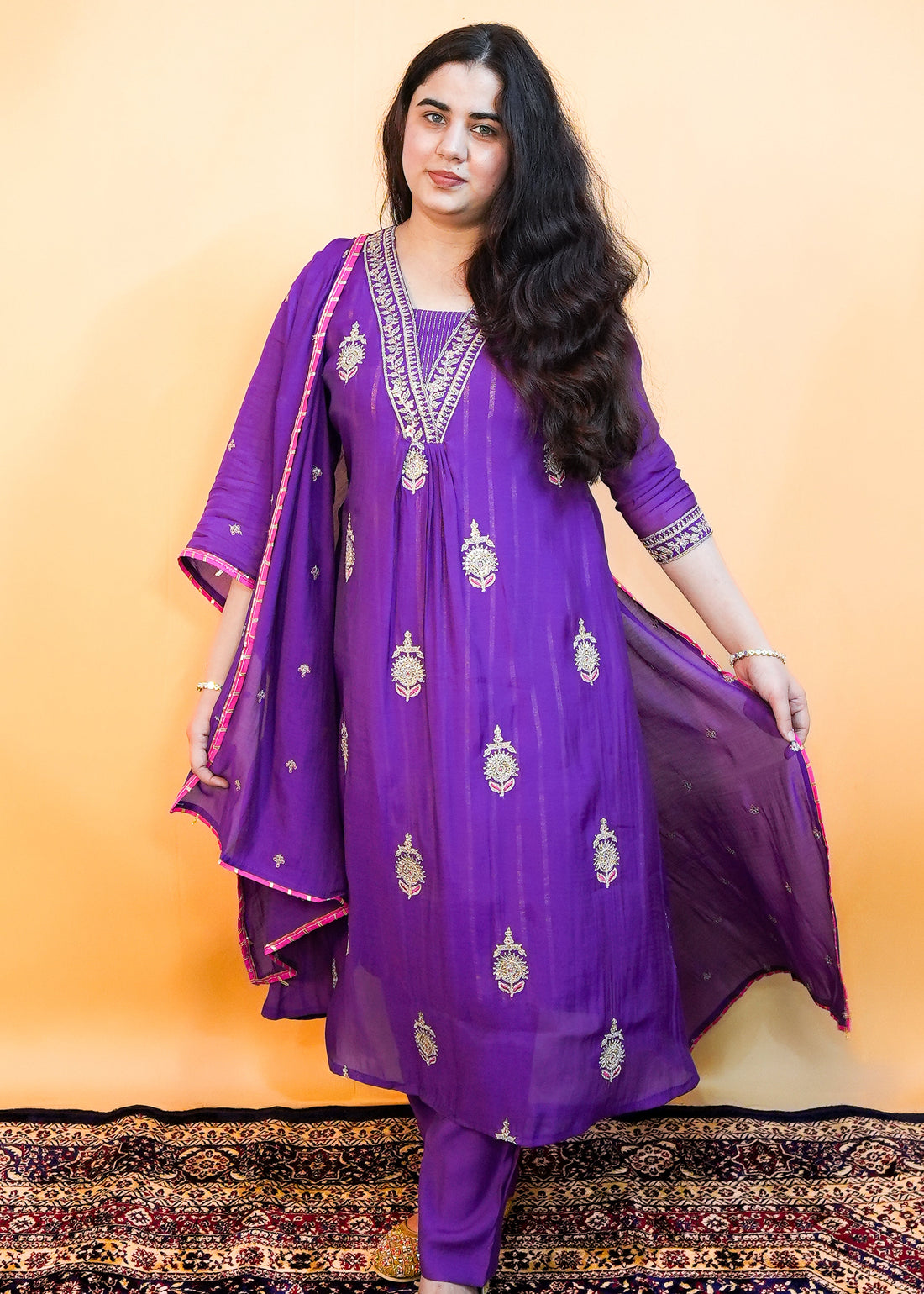 Woman wearing a purple traditional outfit with gold embroidery on a patterned rug.