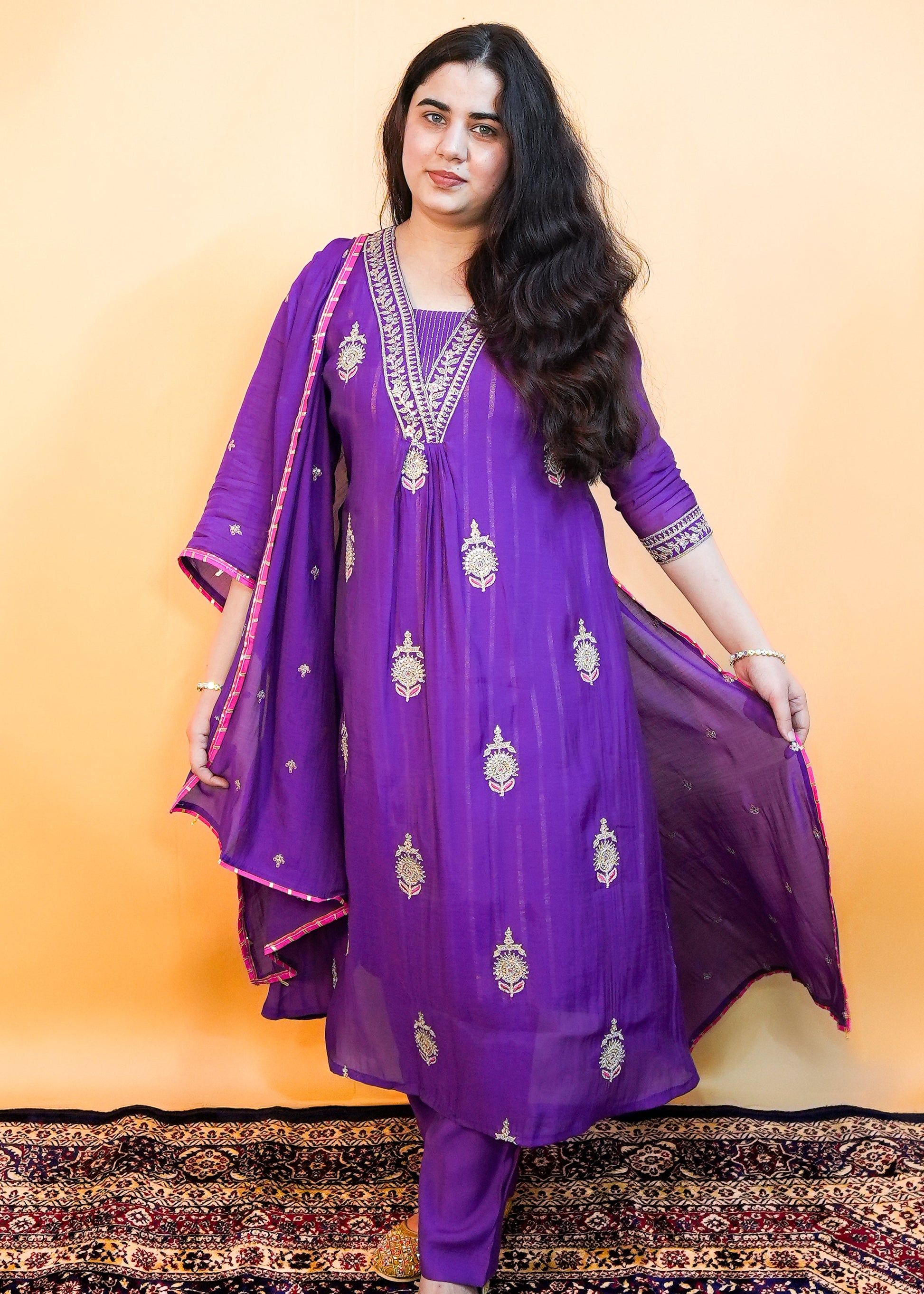 Woman wearing a purple traditional outfit with gold embroidery on a patterned rug.