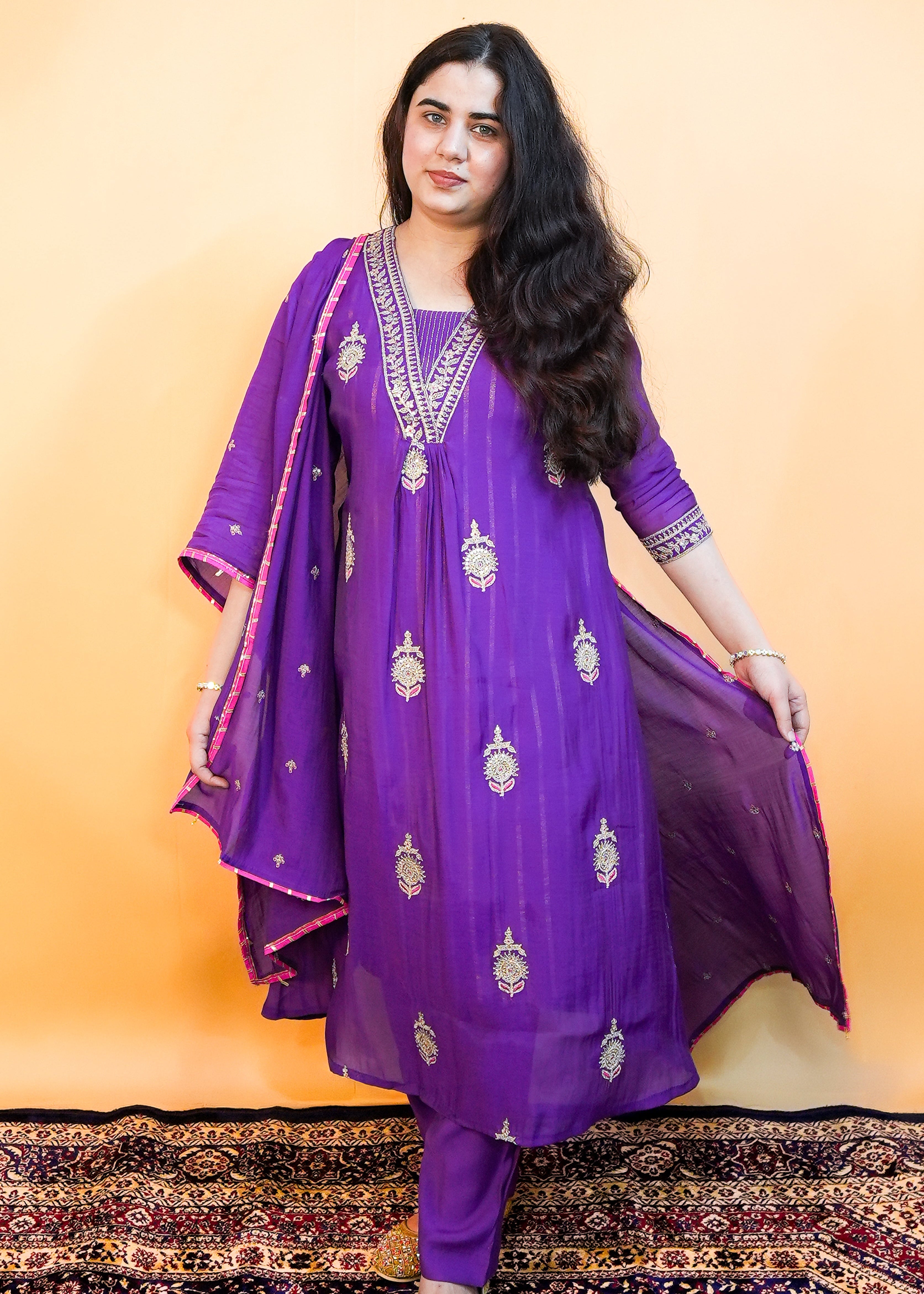 Woman wearing a purple traditional outfit with gold embroidery on a patterned rug.