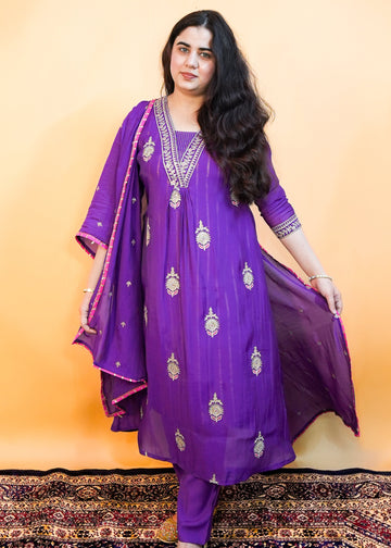 Woman wearing a purple traditional outfit with gold embroidery on a patterned rug.