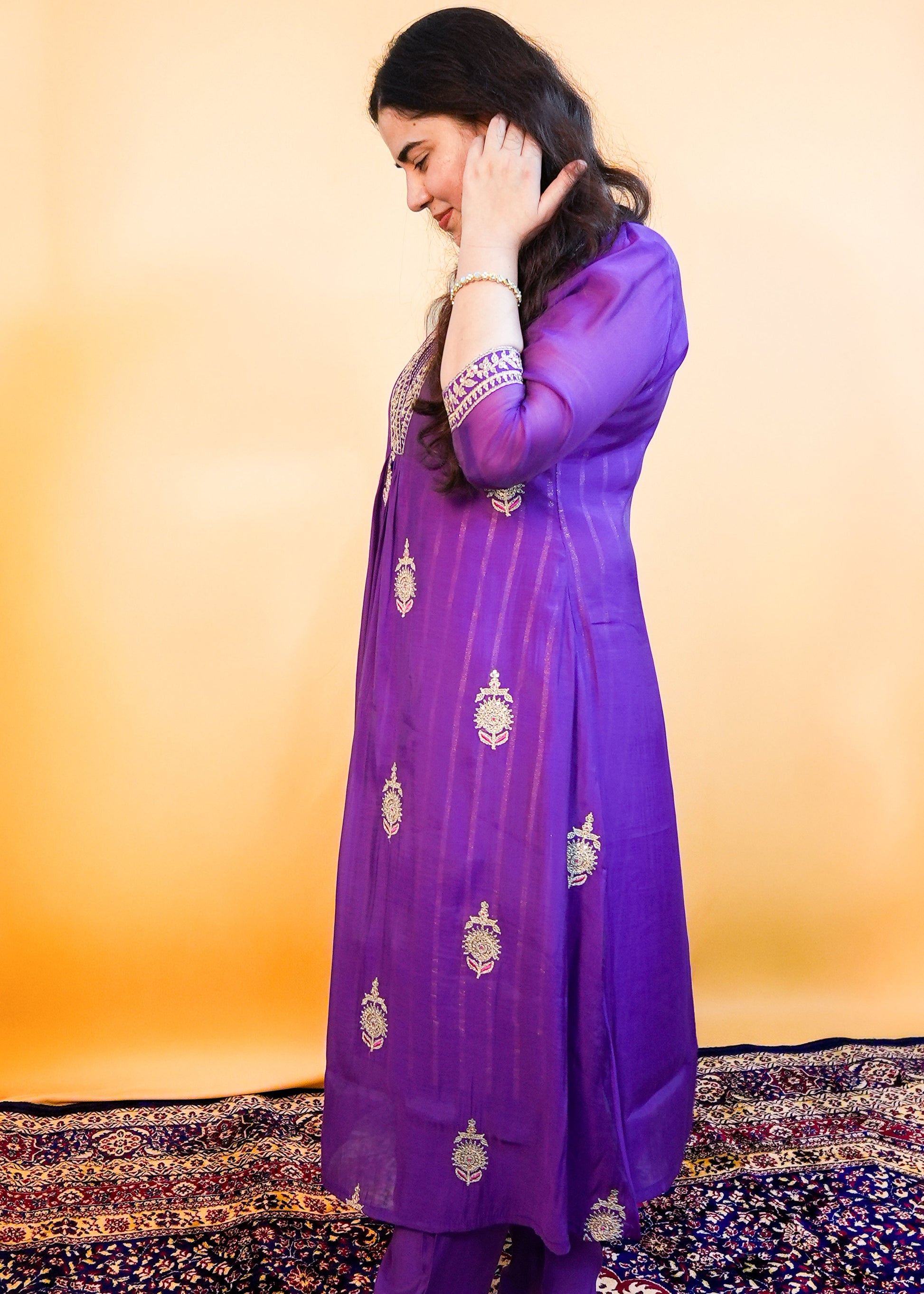 Woman wearing a purple traditional outfit with gold embroidery on a patterned rug.