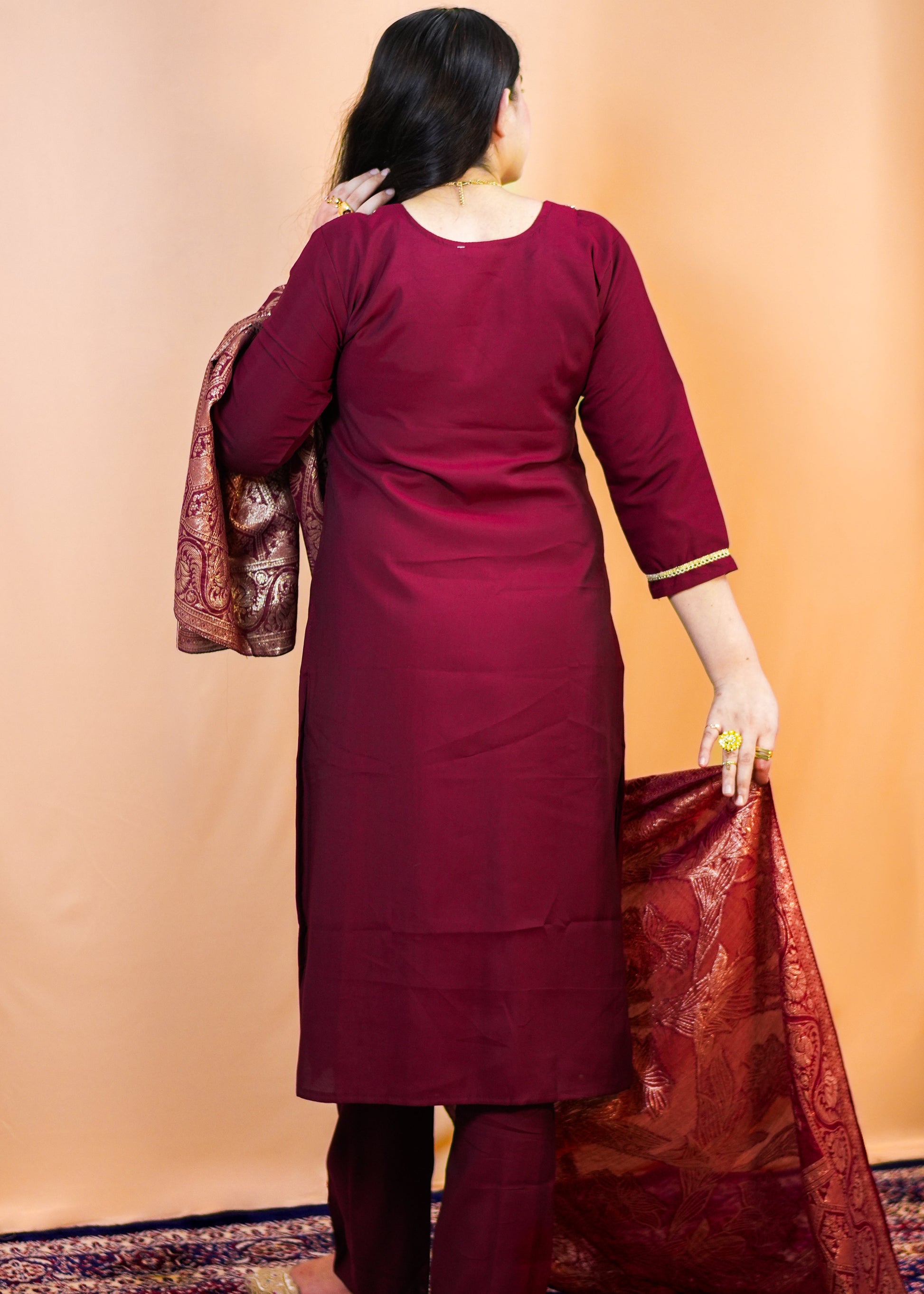 Woman in a maroon traditional outfit holding a red and gold dupatta against a beige wall.