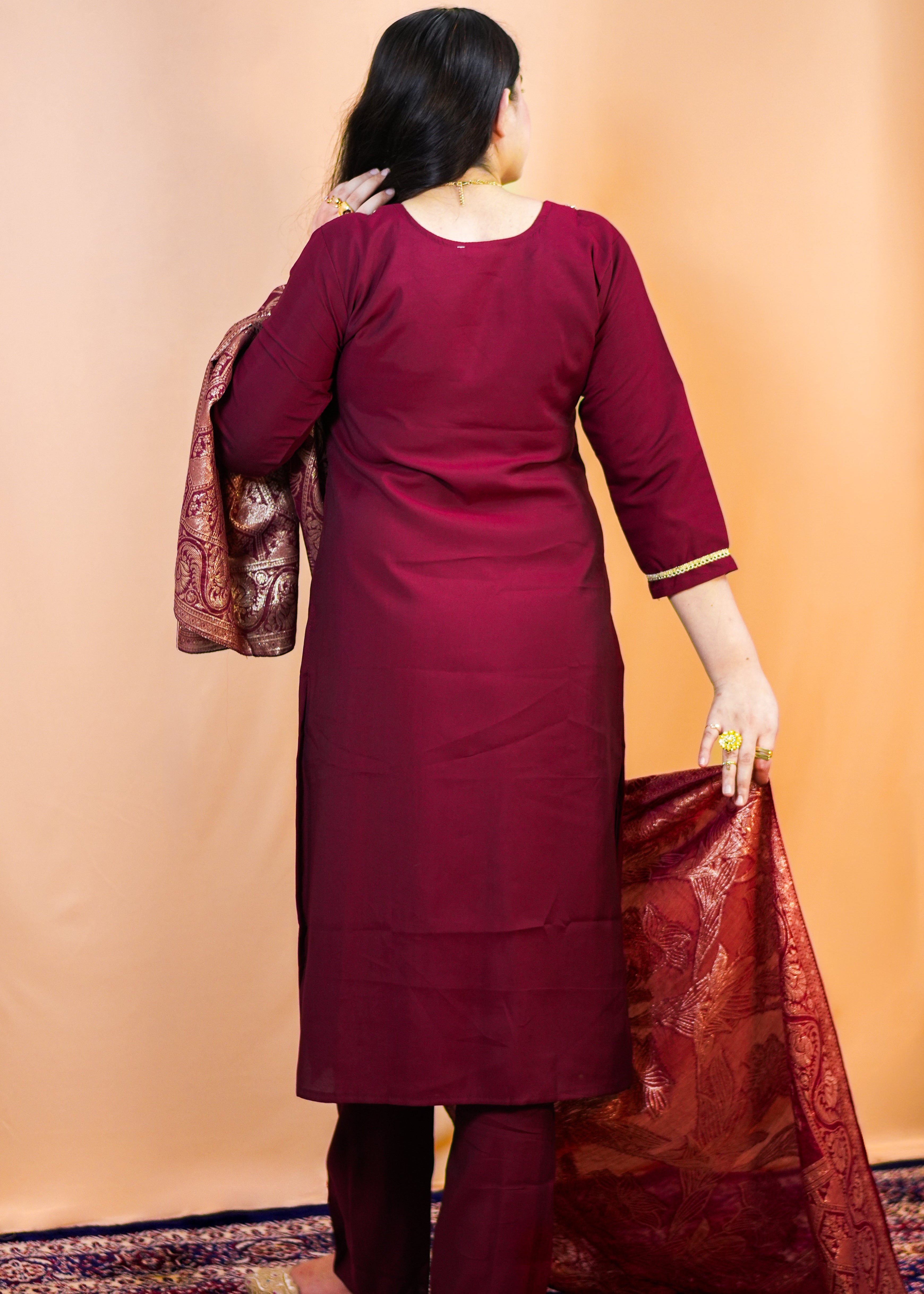 Woman in a maroon traditional outfit holding a red and gold dupatta against a beige wall.