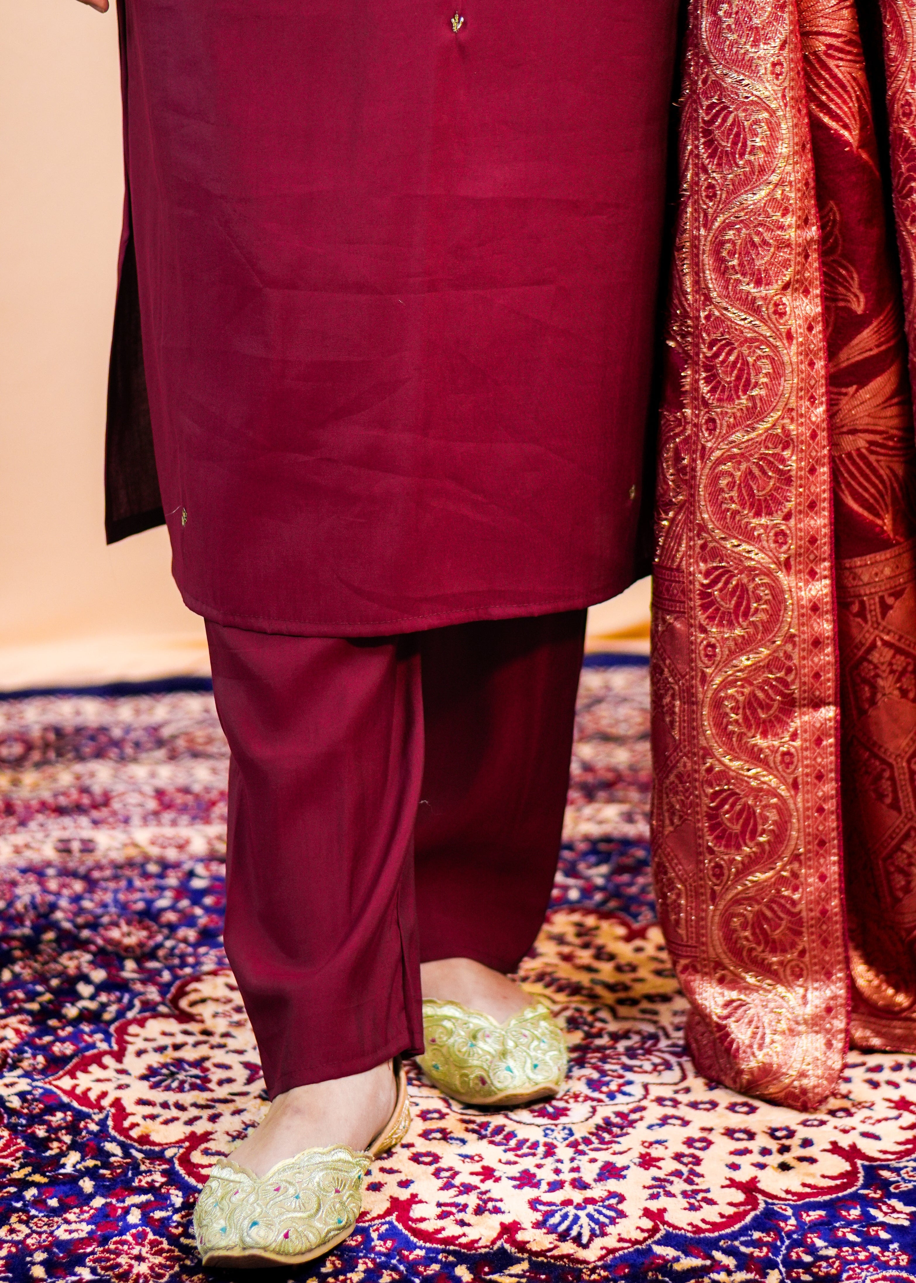 Person wearing a maroon traditional outfit with gold shoes on a patterned rug.