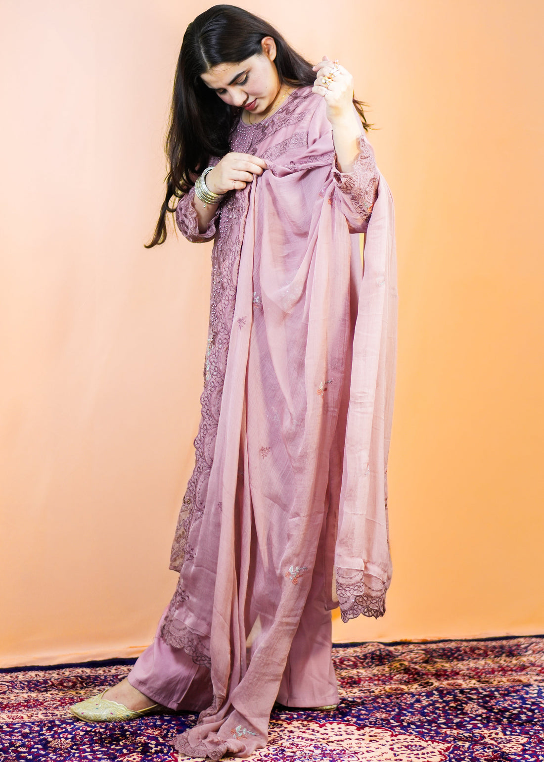 Woman in a traditional outfit with a pinkish-purple dupatta on a patterned rug.