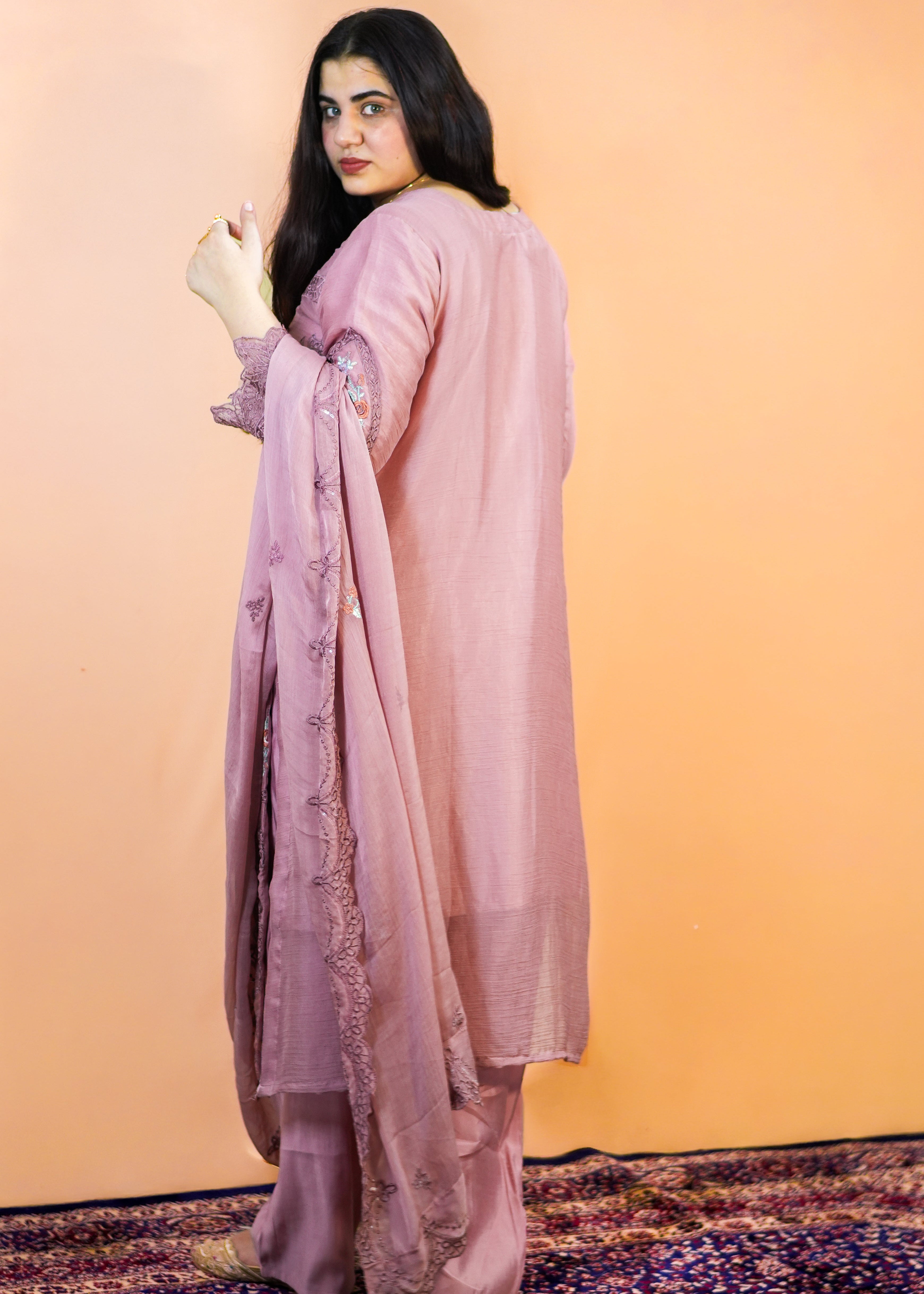Woman in a pink traditional outfit standing on a patterned rug with a peach-colored wall background
