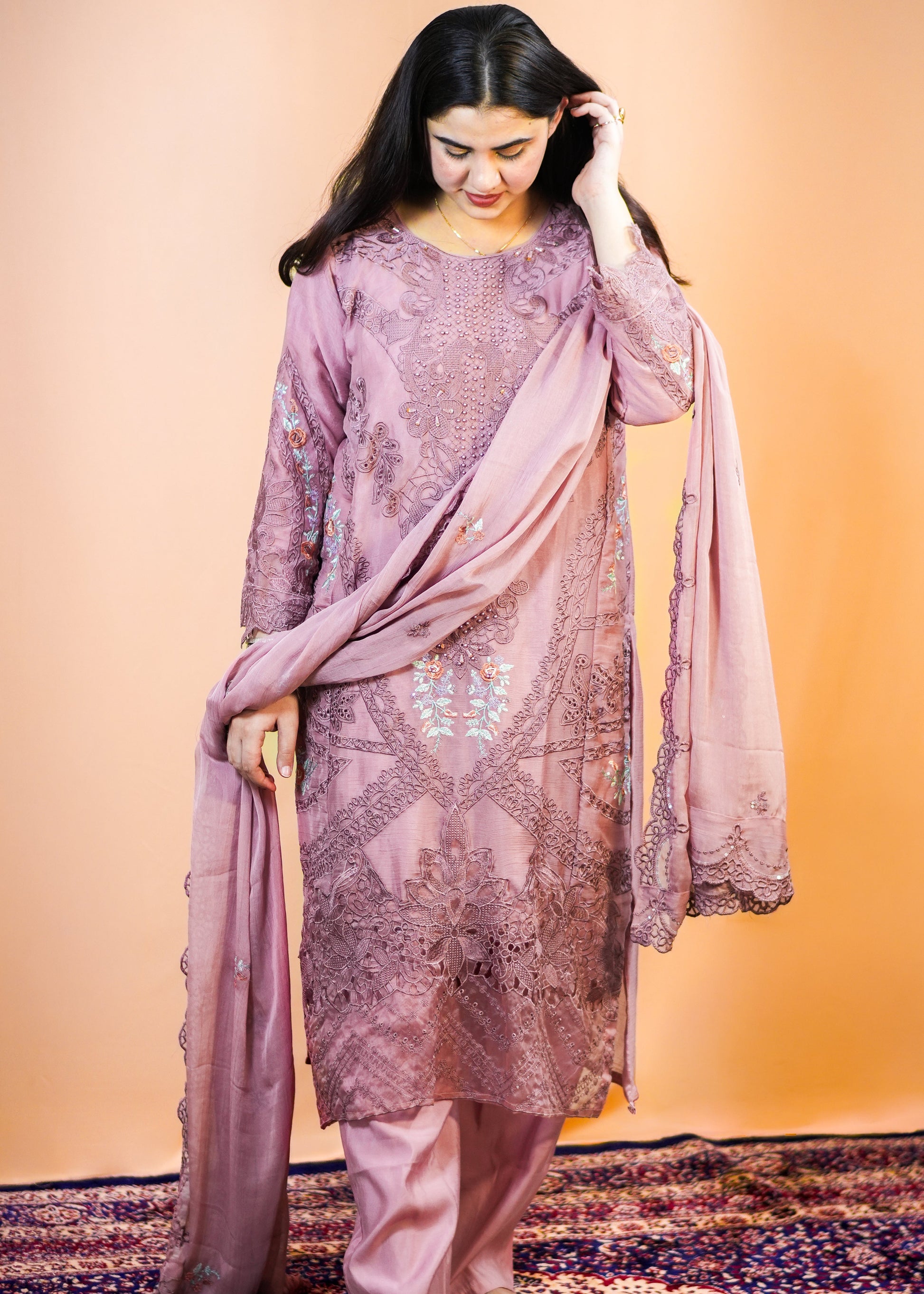 Woman in a traditional outfit with a pinkish-purple dupatta on a patterned rug.