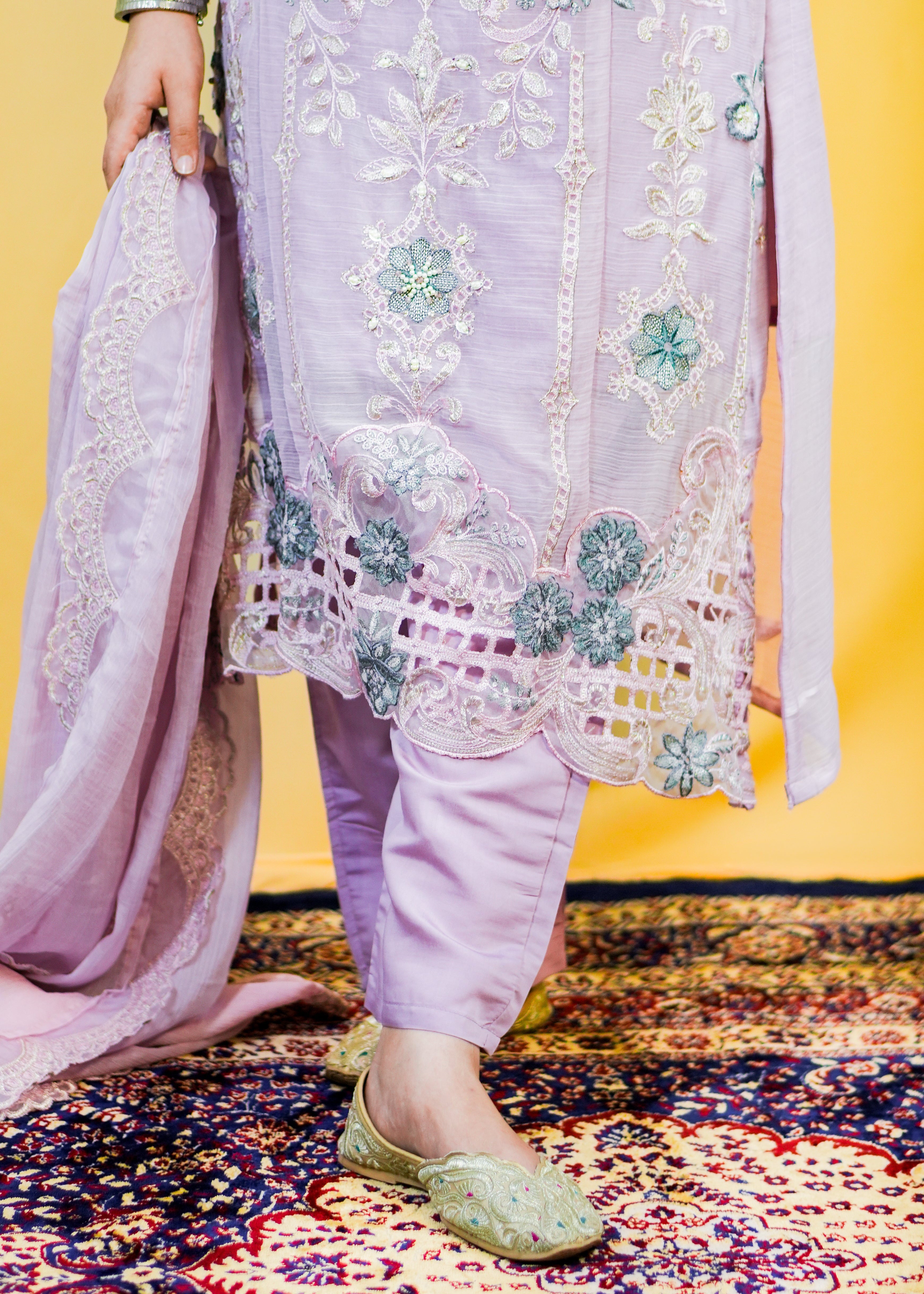 Person wearing a light purple embroidered outfit on a patterned rug with a yellow background