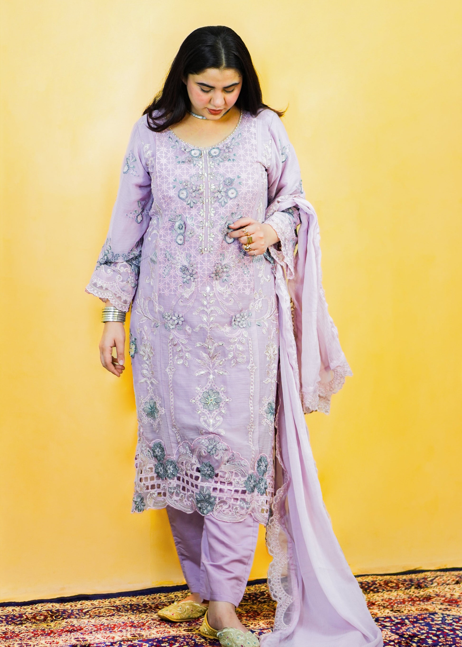Woman in a light purple traditional outfit with floral patterns against a yellow wall.