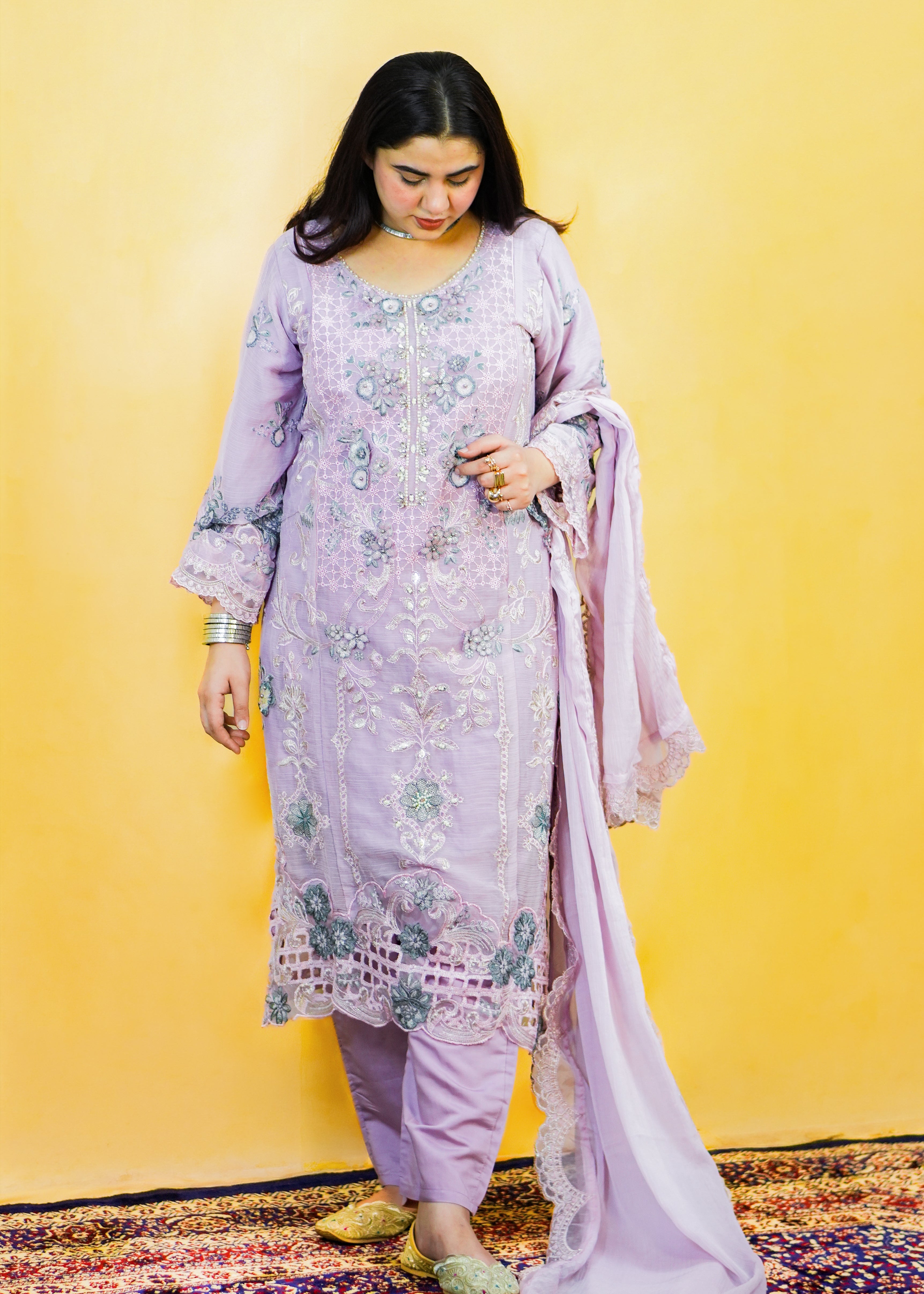 Woman in a light purple traditional outfit with floral patterns against a yellow wall.