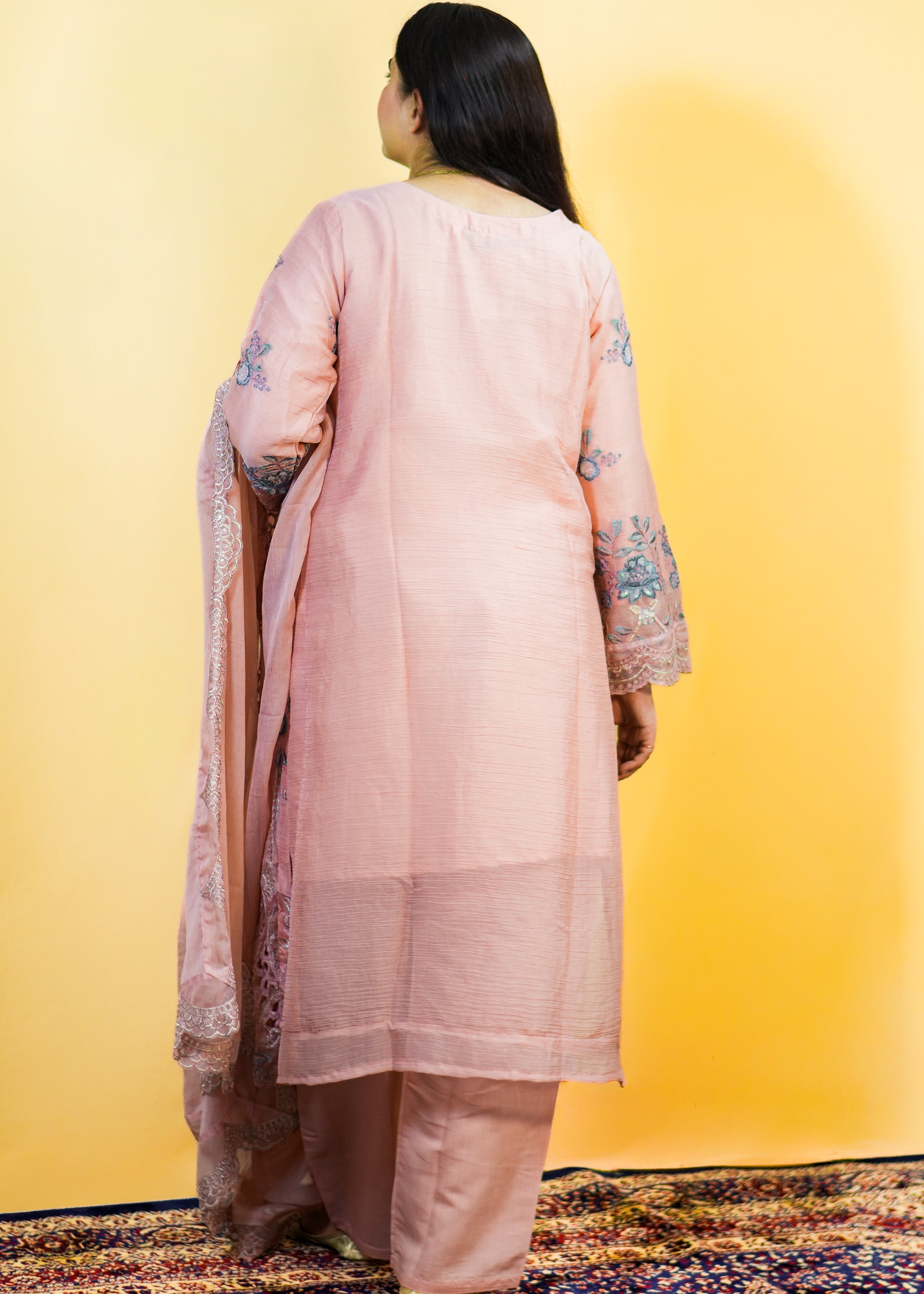 Woman in a light pink traditional outfit standing on a patterned rug with a yellow wall background