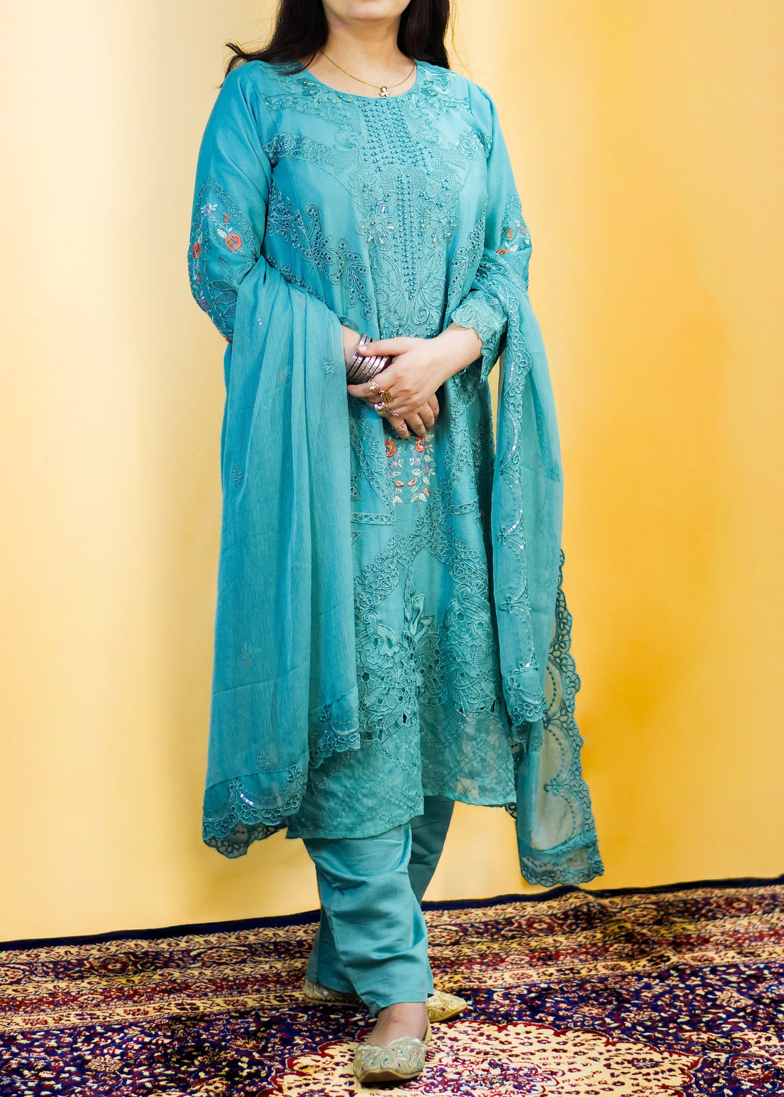 Woman wearing a light blue embroidered traditional outfit on a patterned rug with a yellow background
