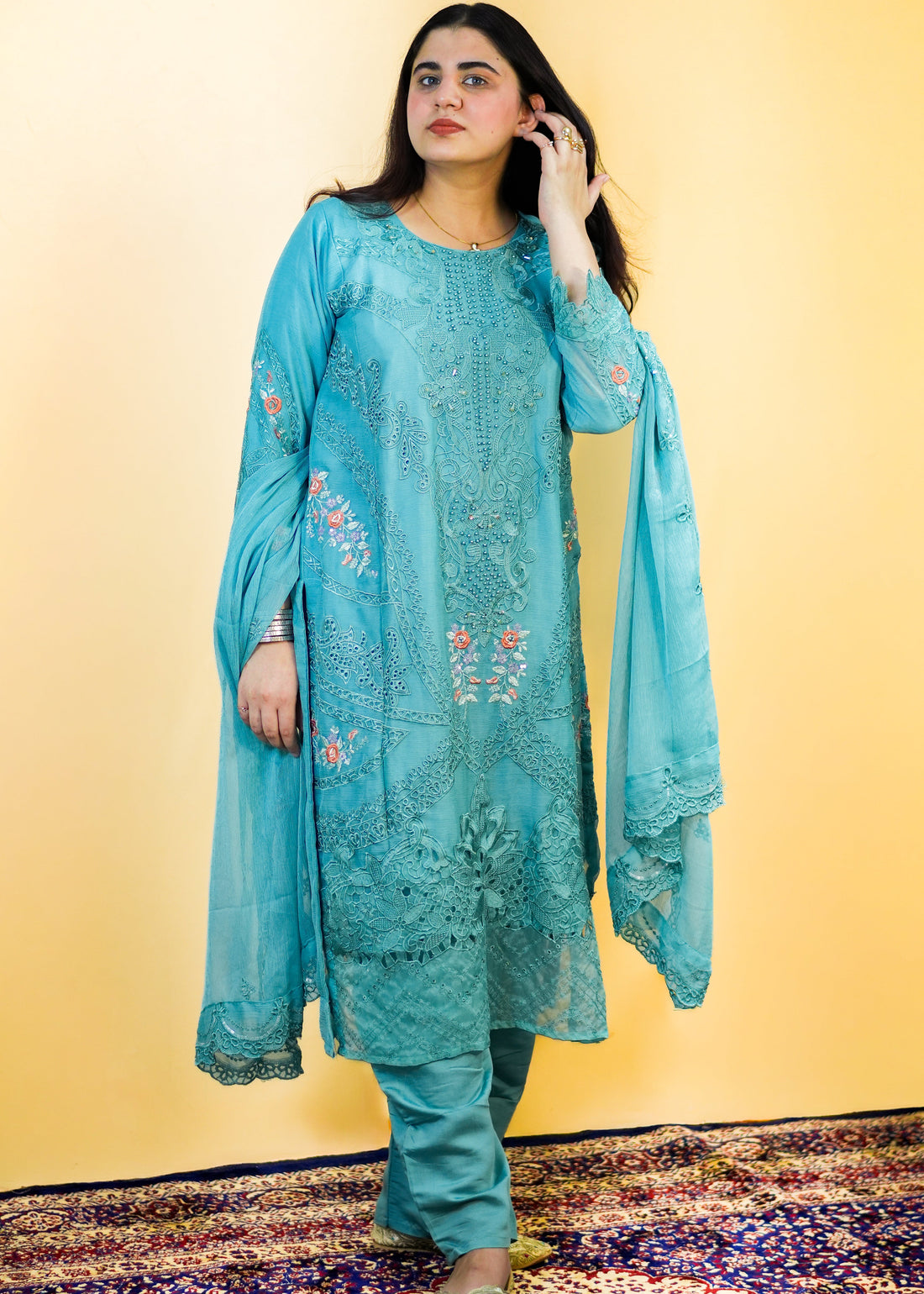 Woman wearing a light blue embroidered traditional outfit on a patterned rug with a yellow background