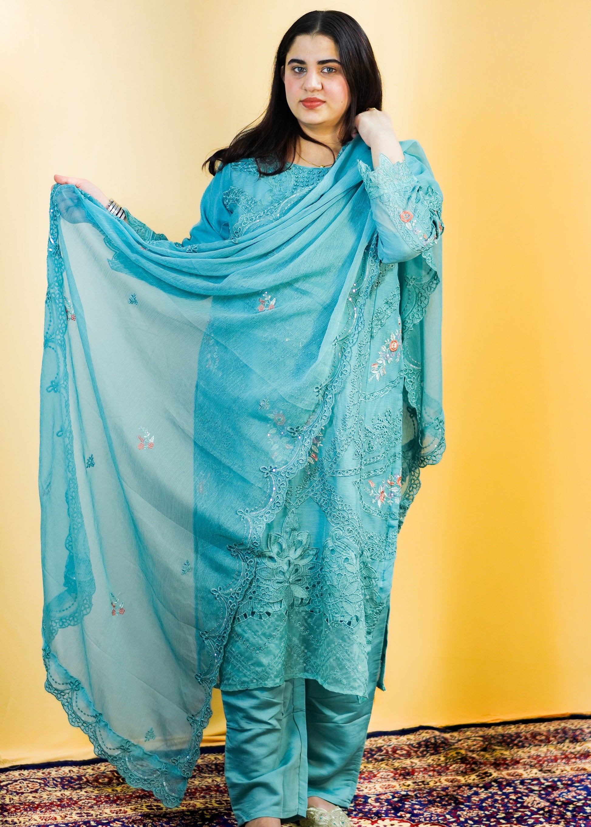 Woman in a blue traditional outfit standing on a patterned rug with a yellow wall background