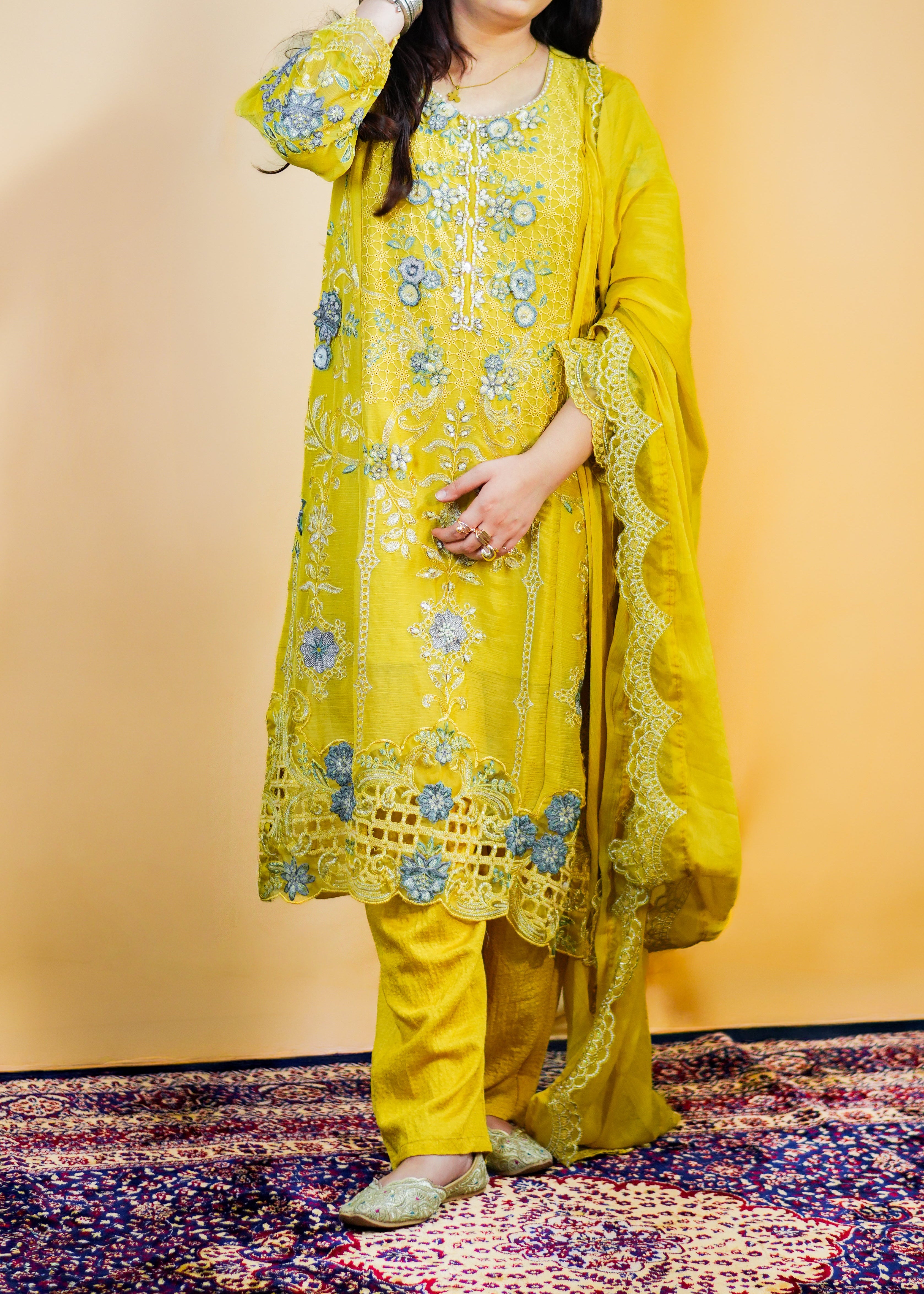 Woman in a yellow traditional outfit with floral patterns on a patterned rug.