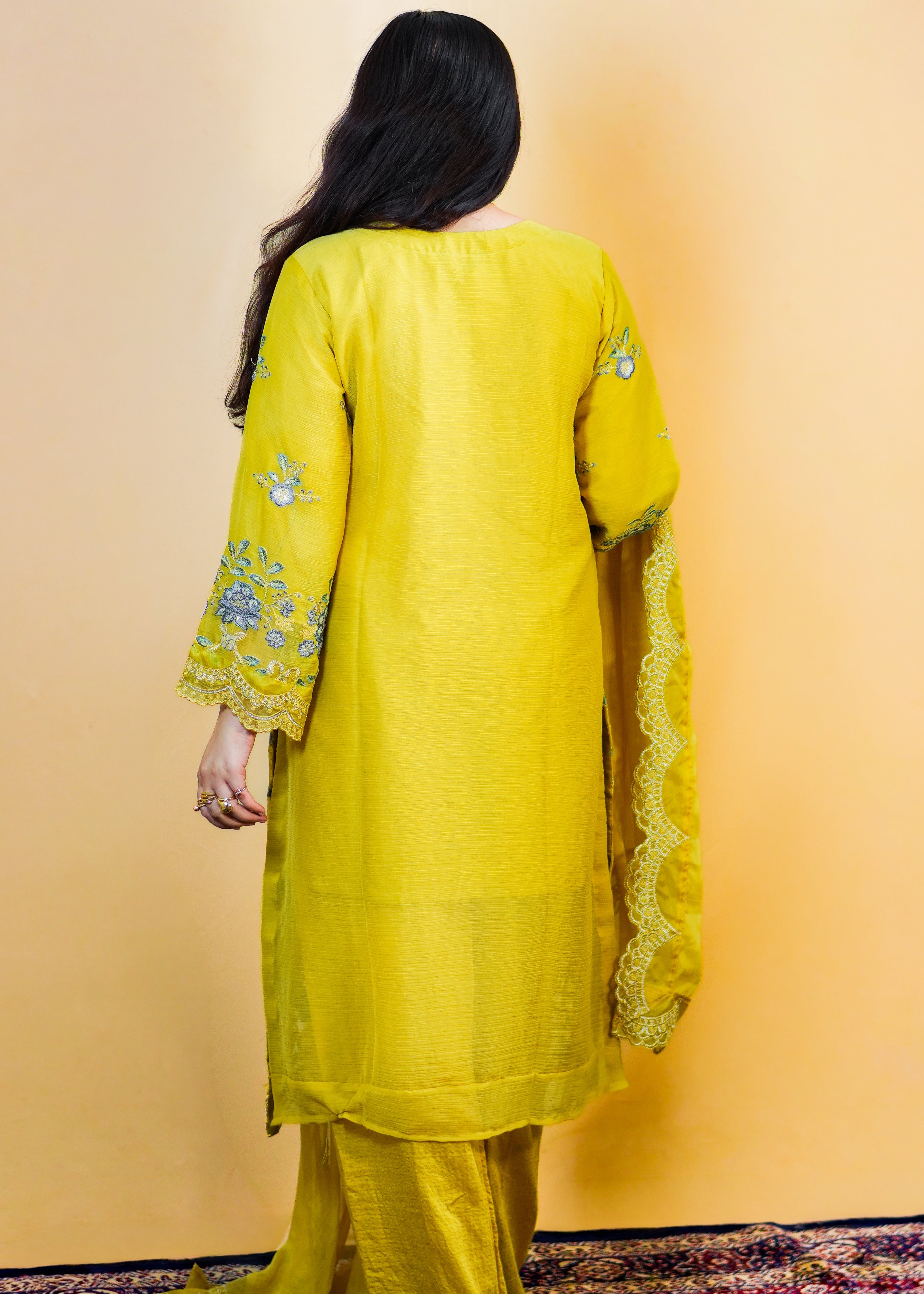 Person wearing a yellow traditional outfit with intricate designs on a beige wall.