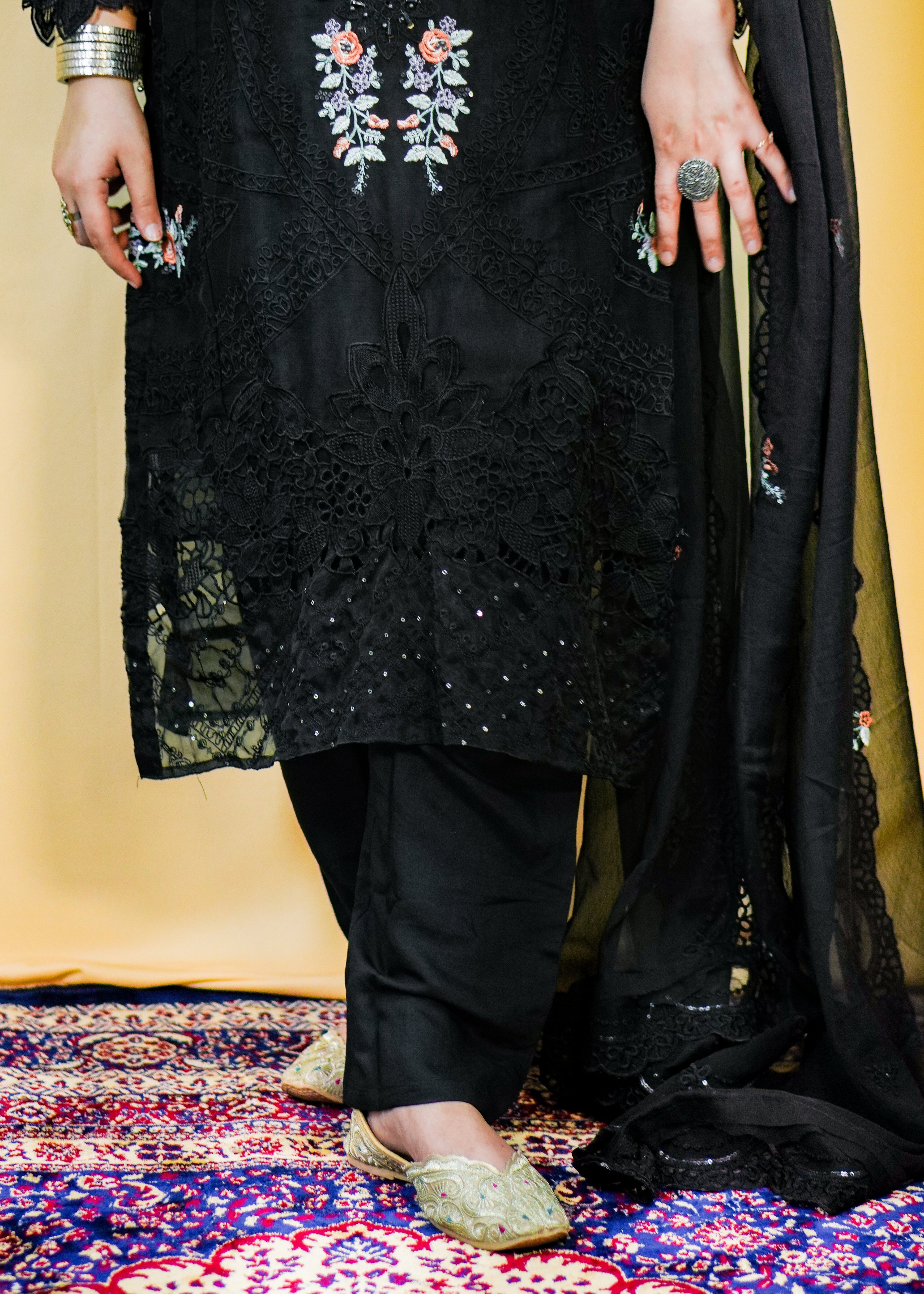 Person wearing a black embroidered outfit standing on a patterned rug.