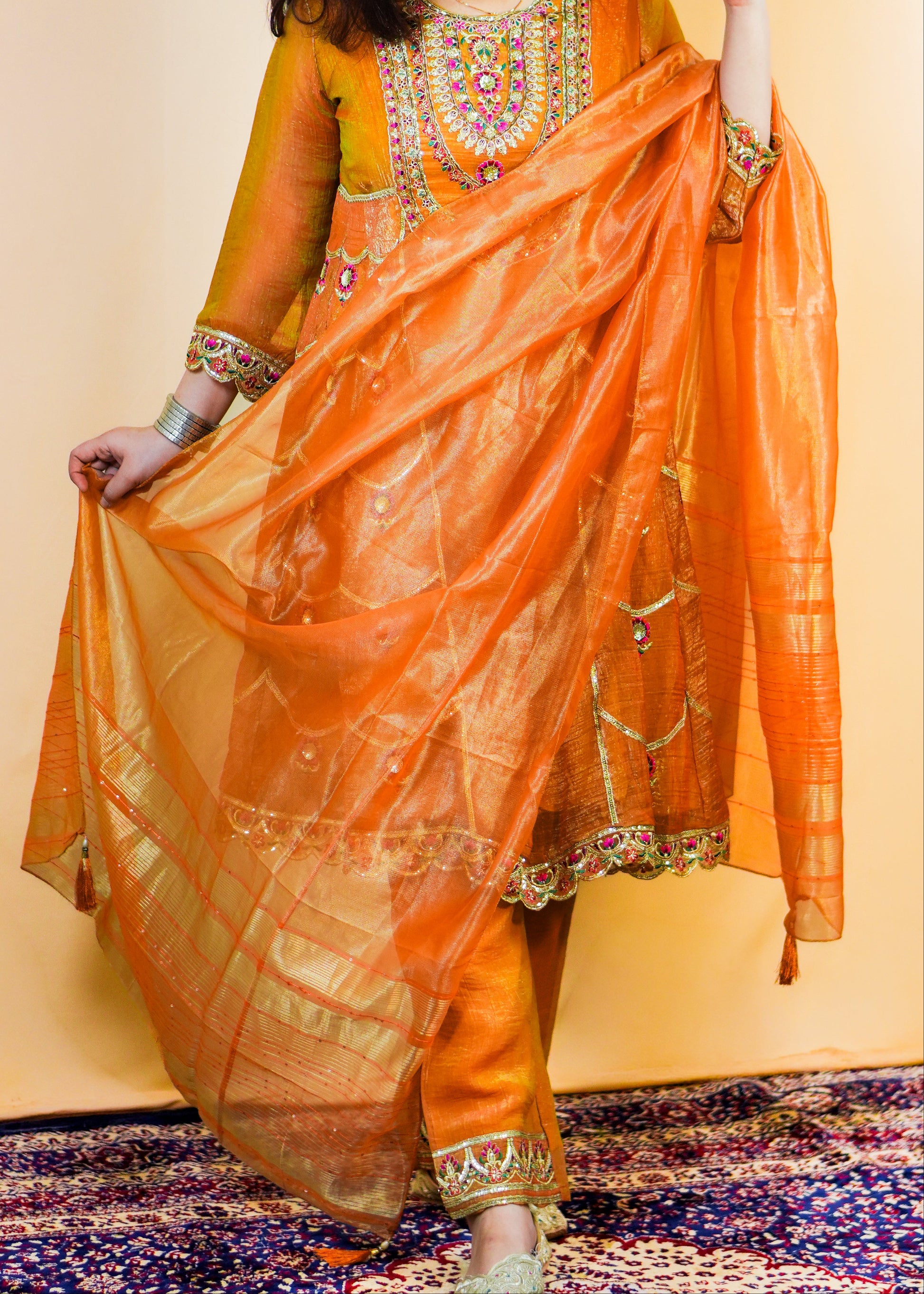 Woman in traditional orange and gold outfit with jewelry, standing on a patterned rug.