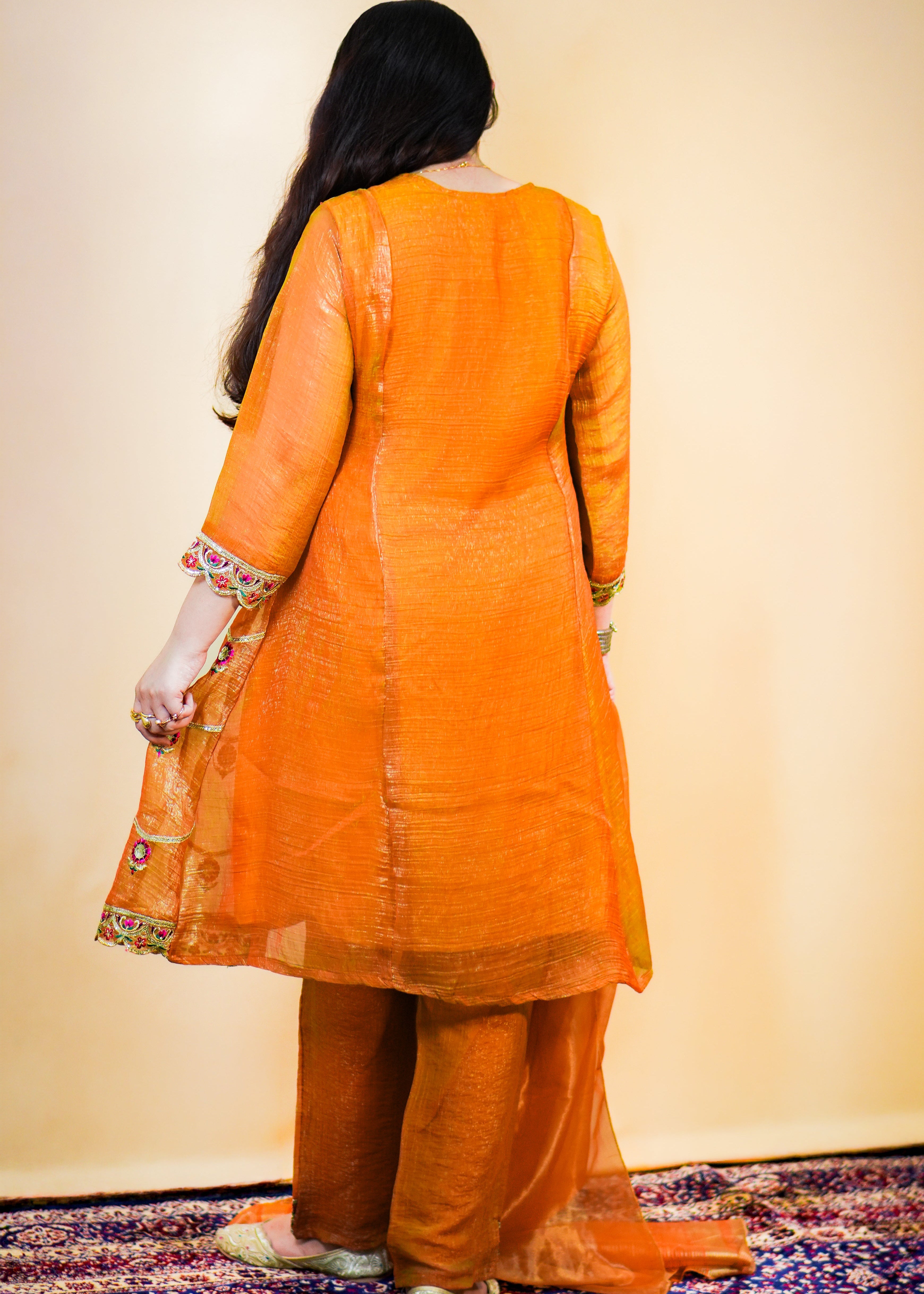 Person wearing an orange traditional outfit standing on a patterned rug.