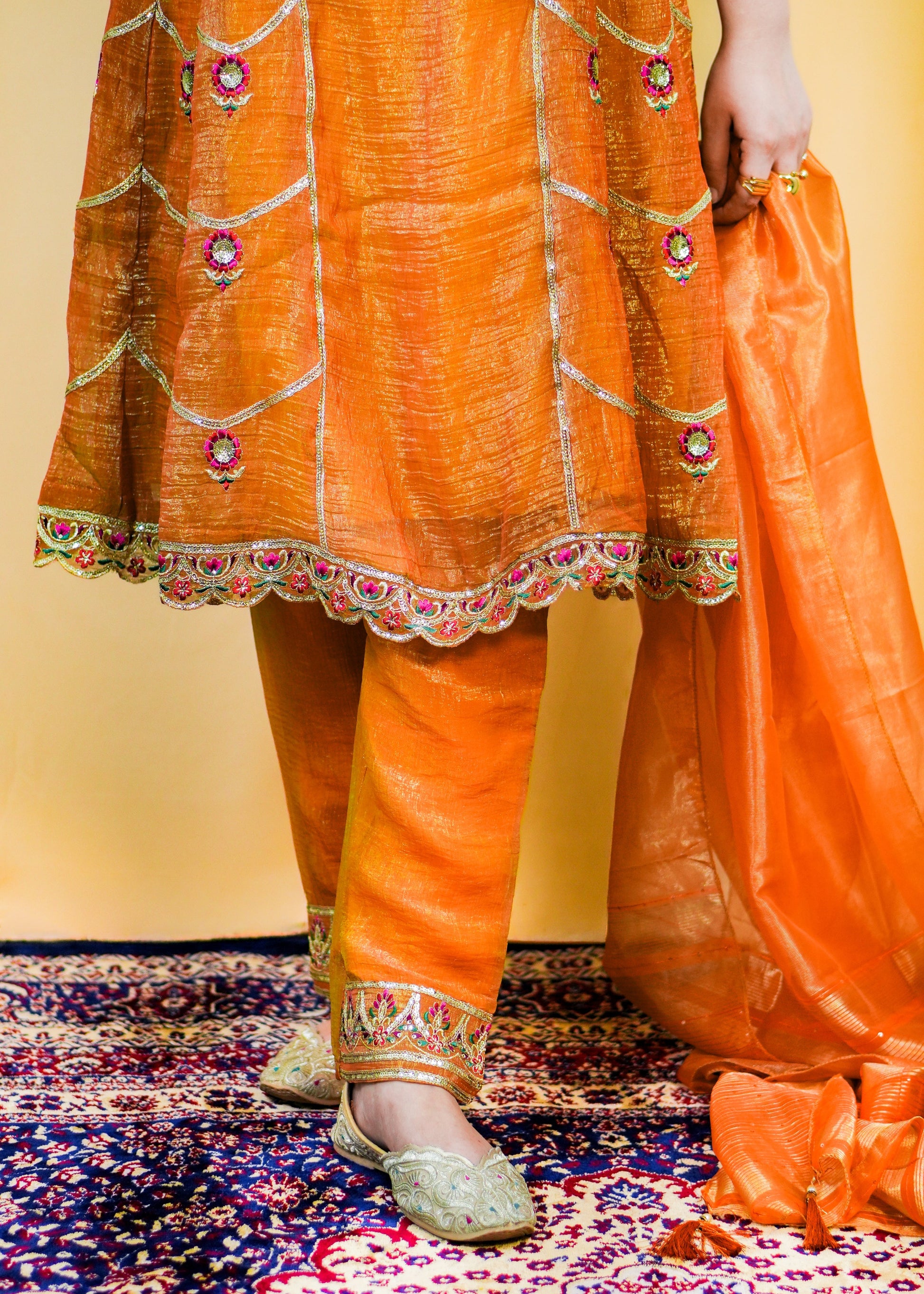 Person wearing an orange traditional outfit with intricate patterns on a patterned rug.