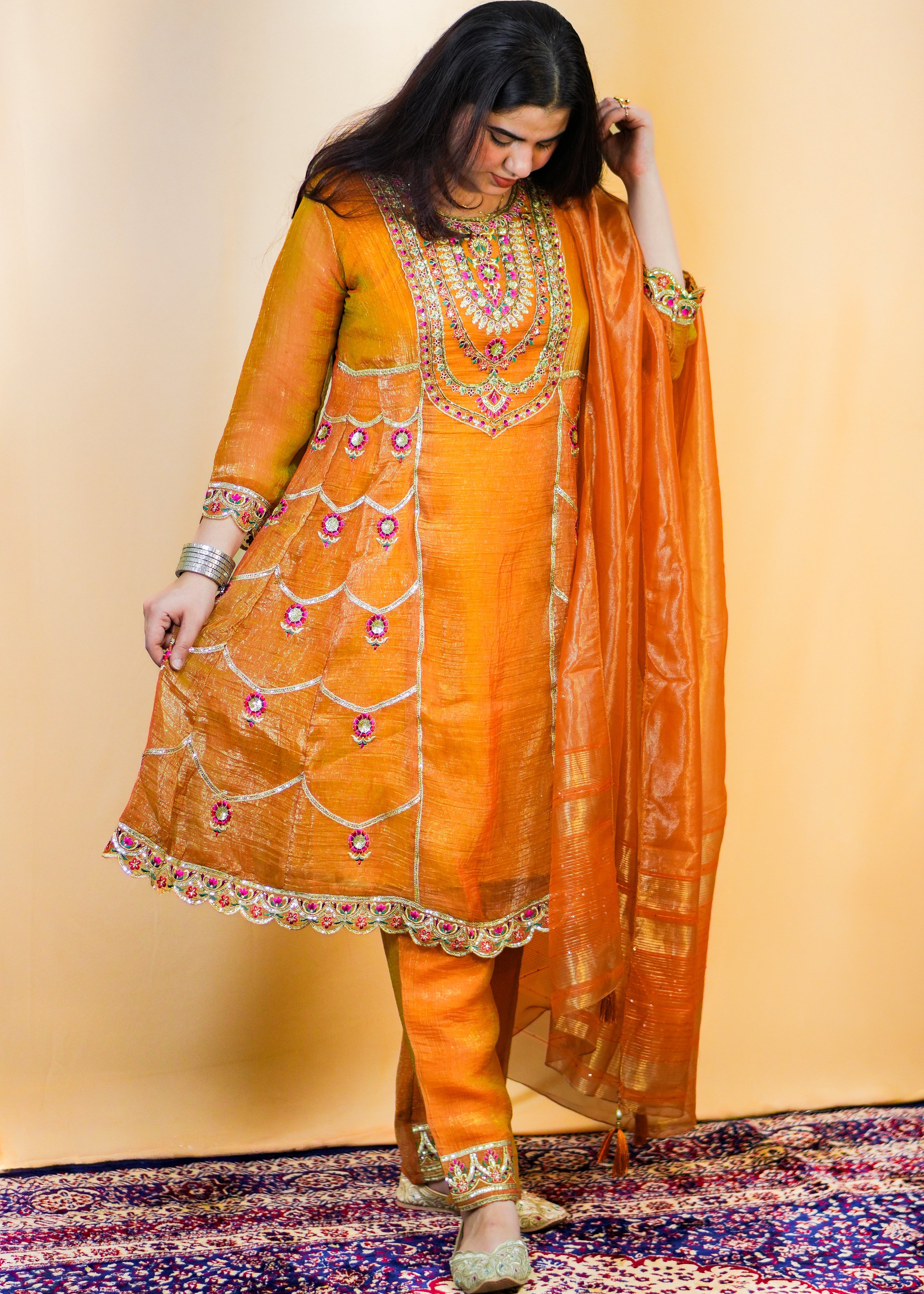 Woman in an orange traditional outfit with a matching dupatta on a patterned rug.