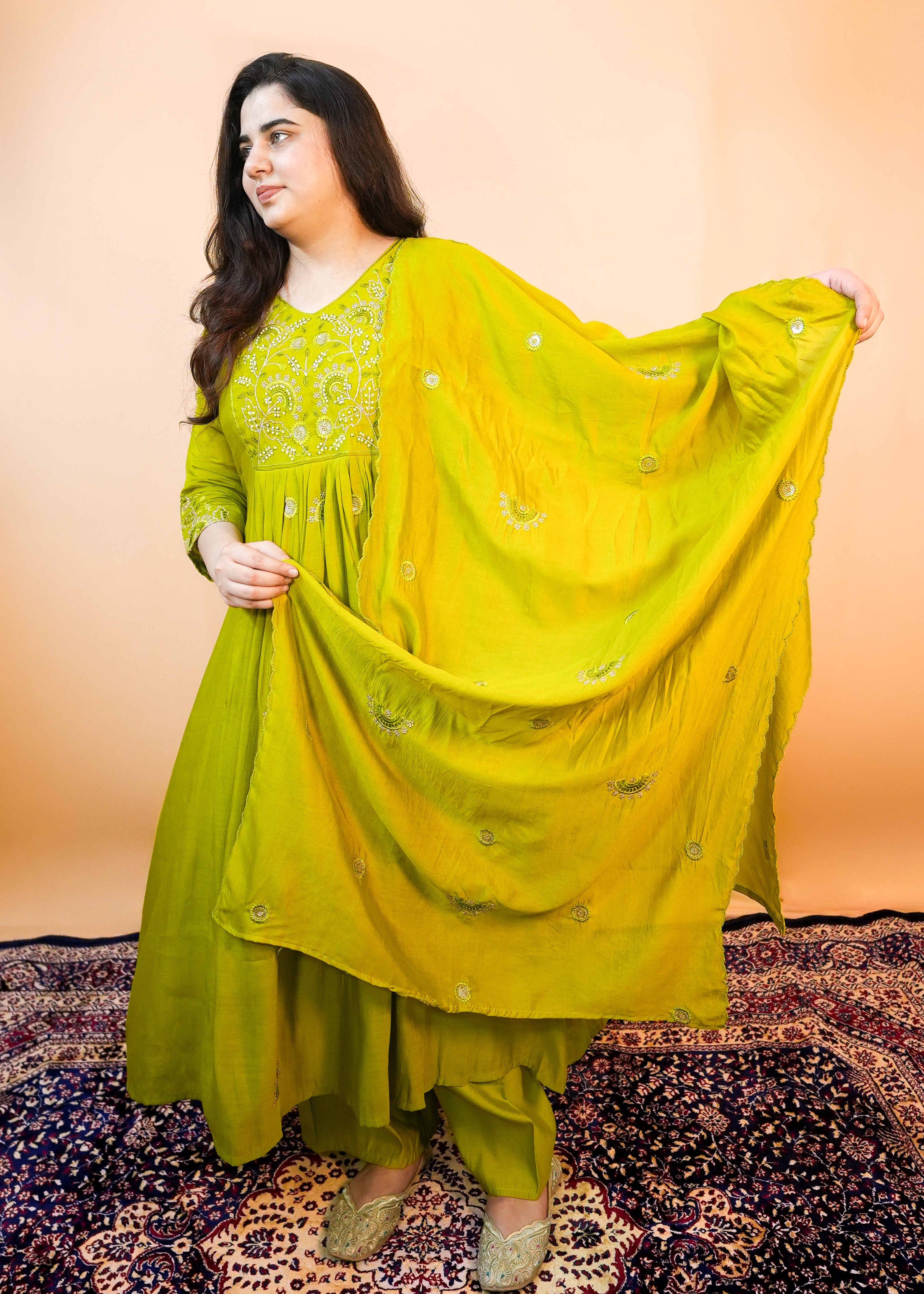 Woman in a bright yellow traditional outfit standing on a patterned rug with an orange wall background.