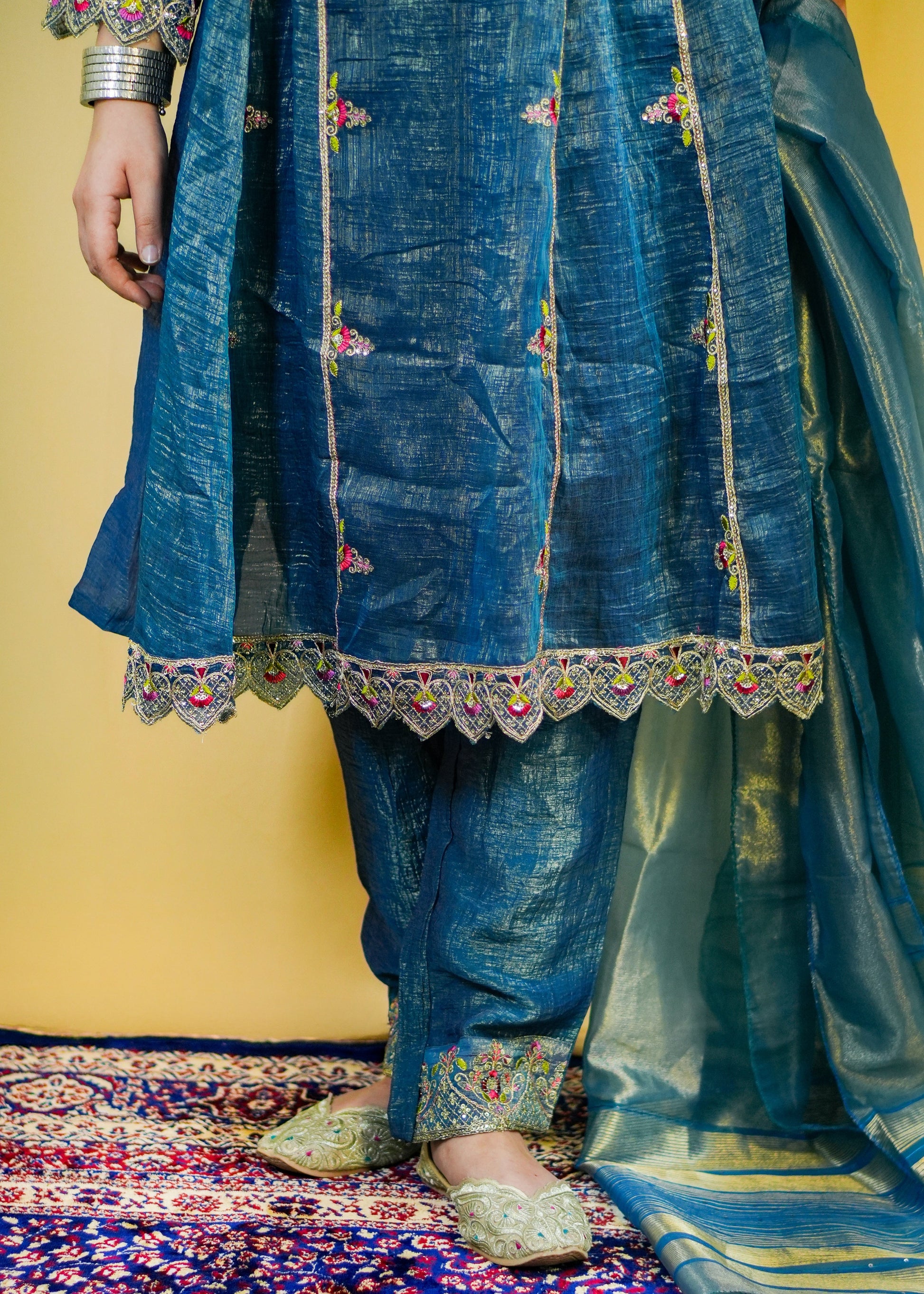 Traditional blue embroidered outfit on a patterned rug with a yellow wall background