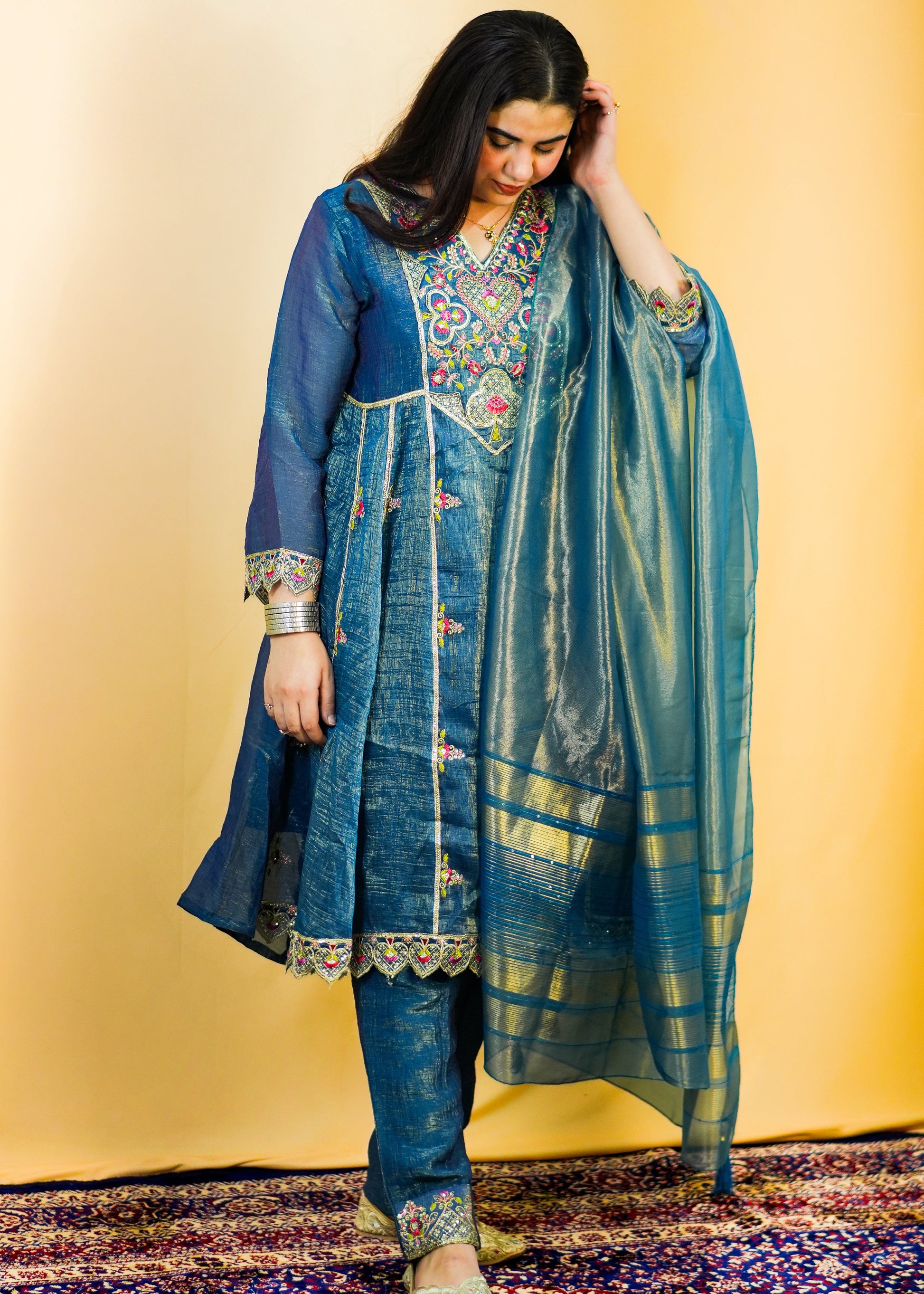 Woman in traditional blue embroidered outfit standing on a patterned rug with a yellow wall background