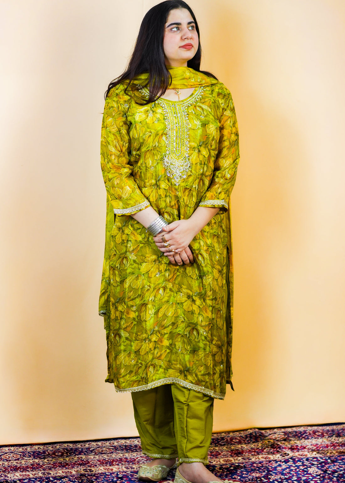 Woman wearing a green floral traditional outfit standing on a patterned rug.