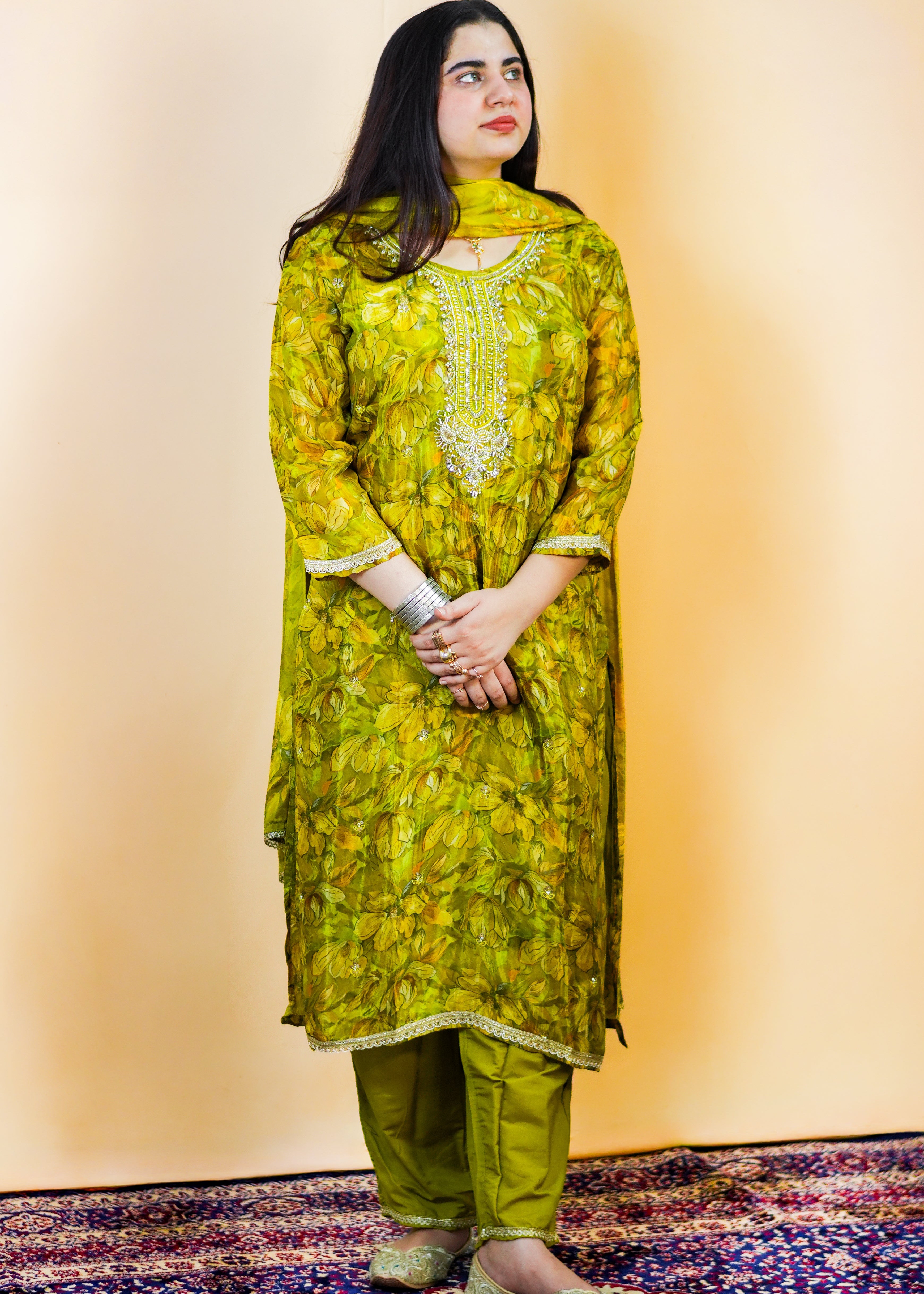 Woman wearing a green floral traditional outfit standing on a patterned rug.