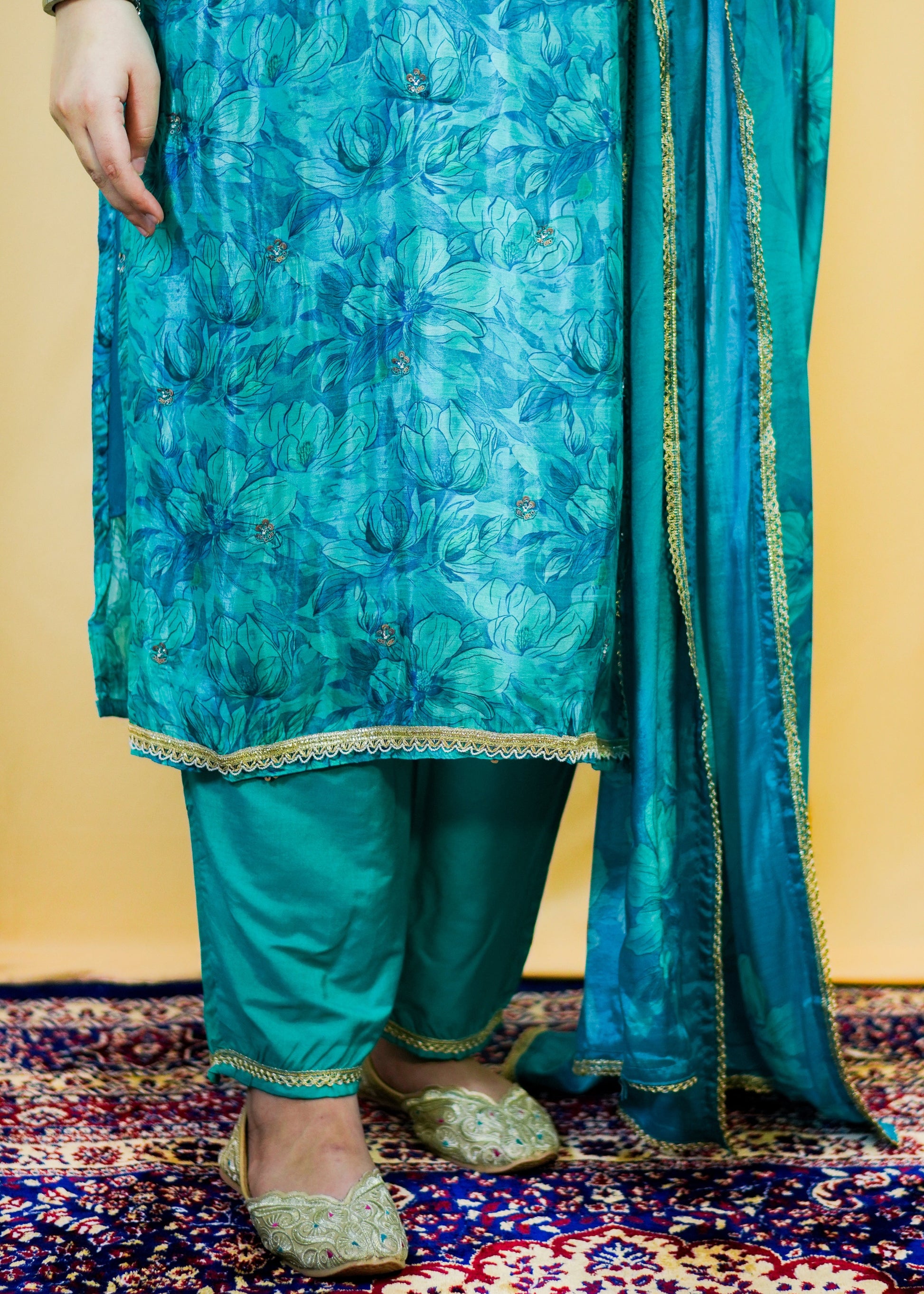 Person wearing a turquoise traditional outfit with a matching dupatta on a patterned rug.