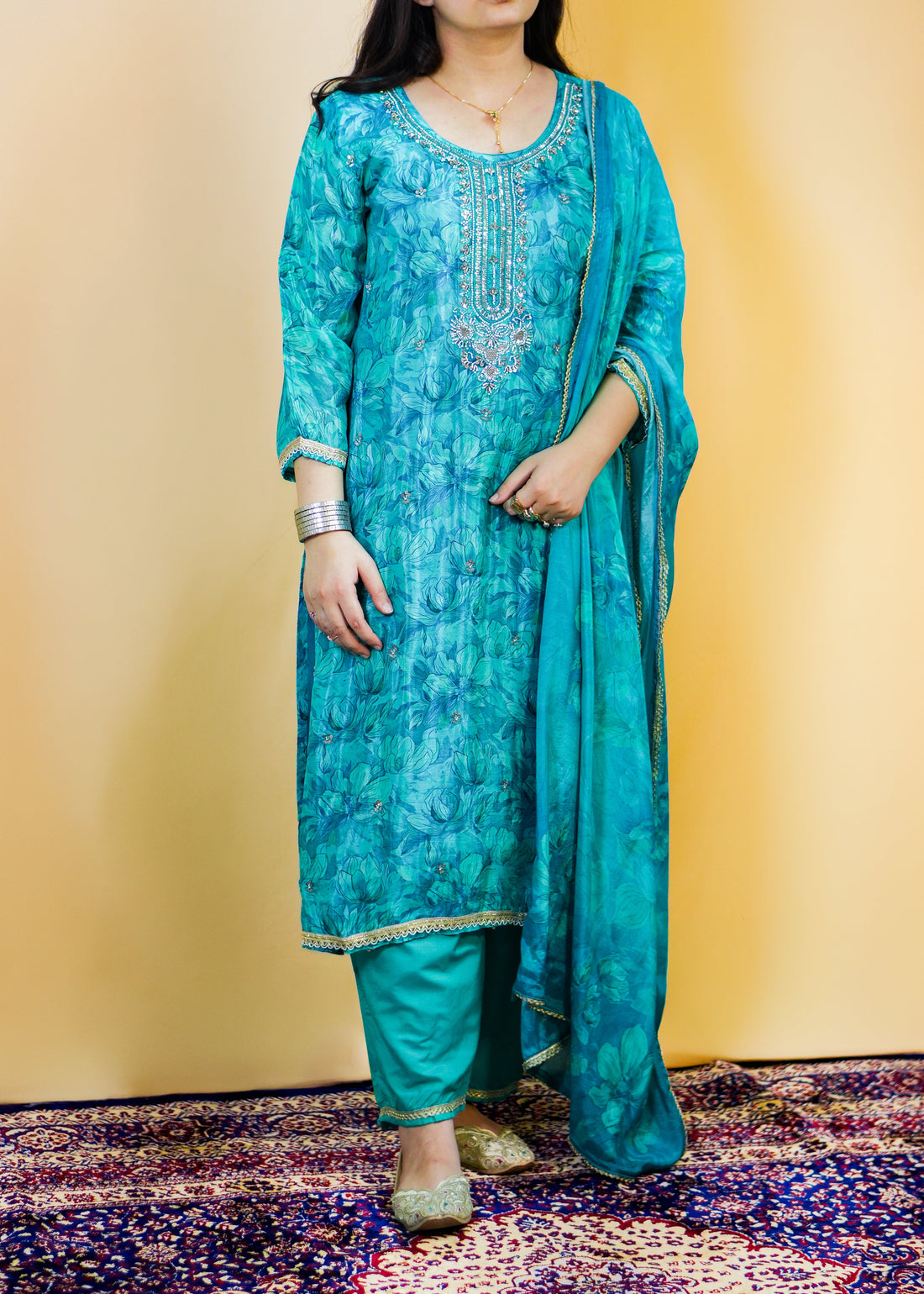 Woman in a blue traditional outfit standing on a patterned rug with a yellow wall background