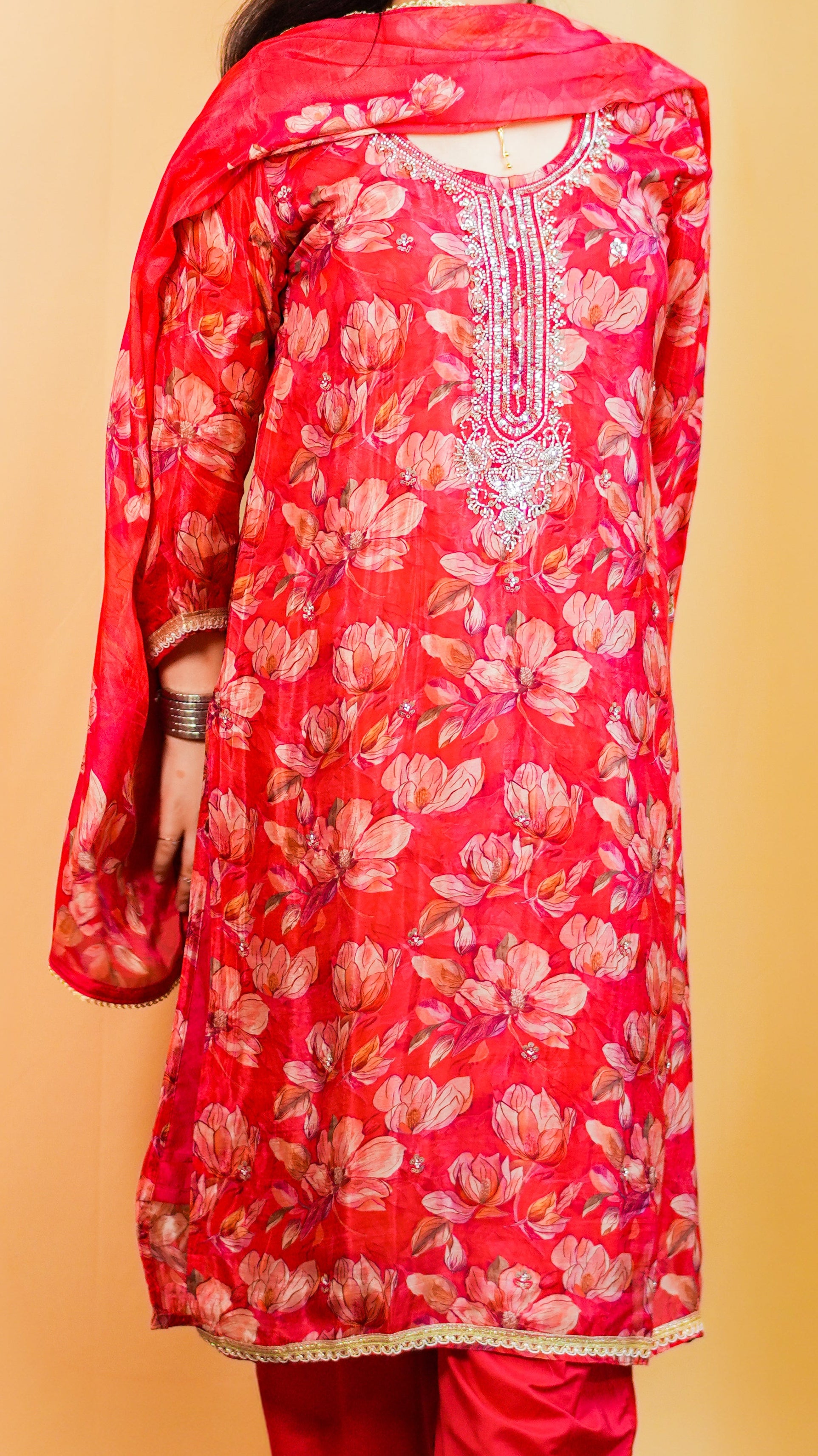 Woman wearing a red floral traditional outfit standing on a patterned rug against a beige wall.