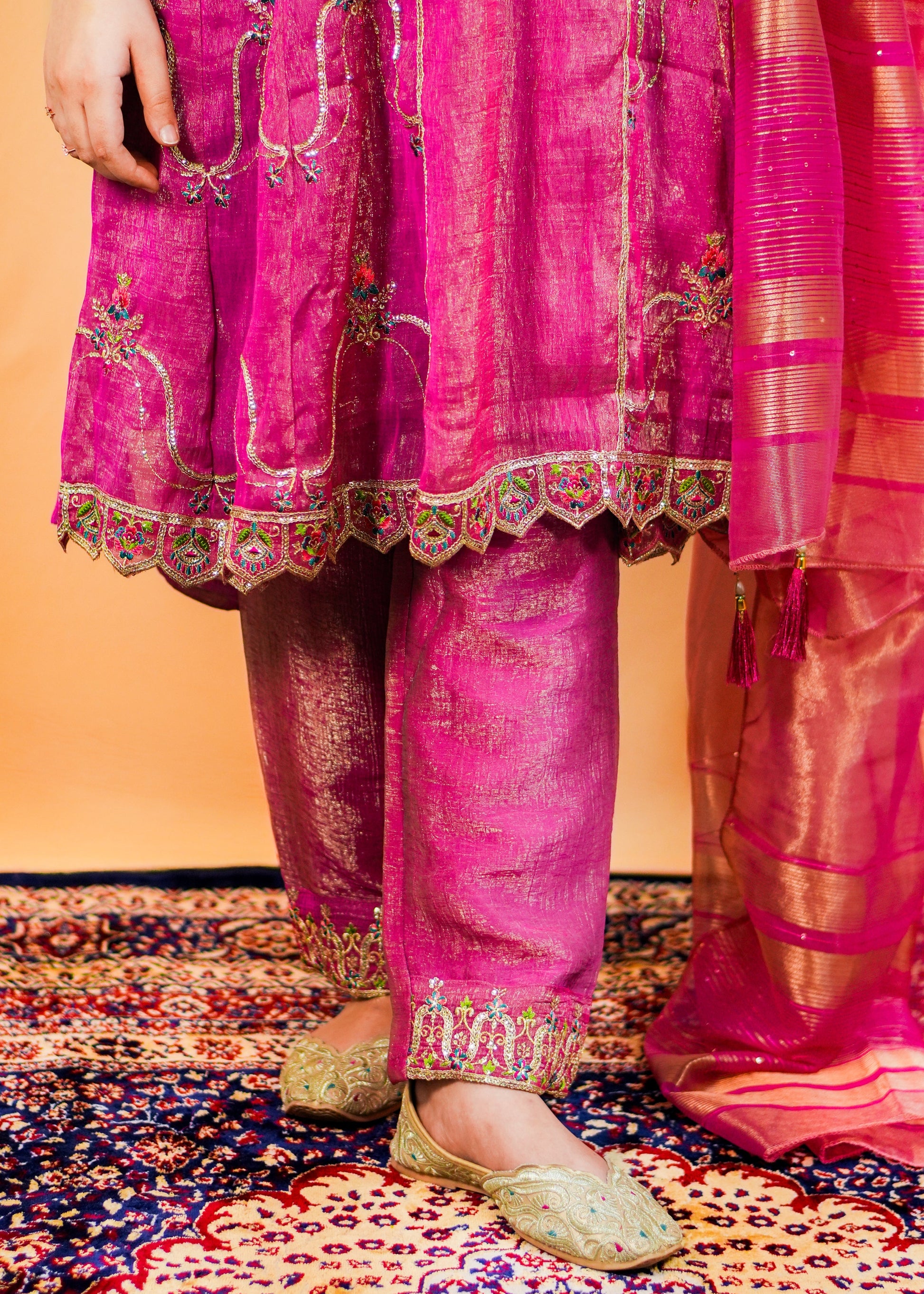 Person wearing a pink traditional outfit with intricate embroidery on a patterned carpet.